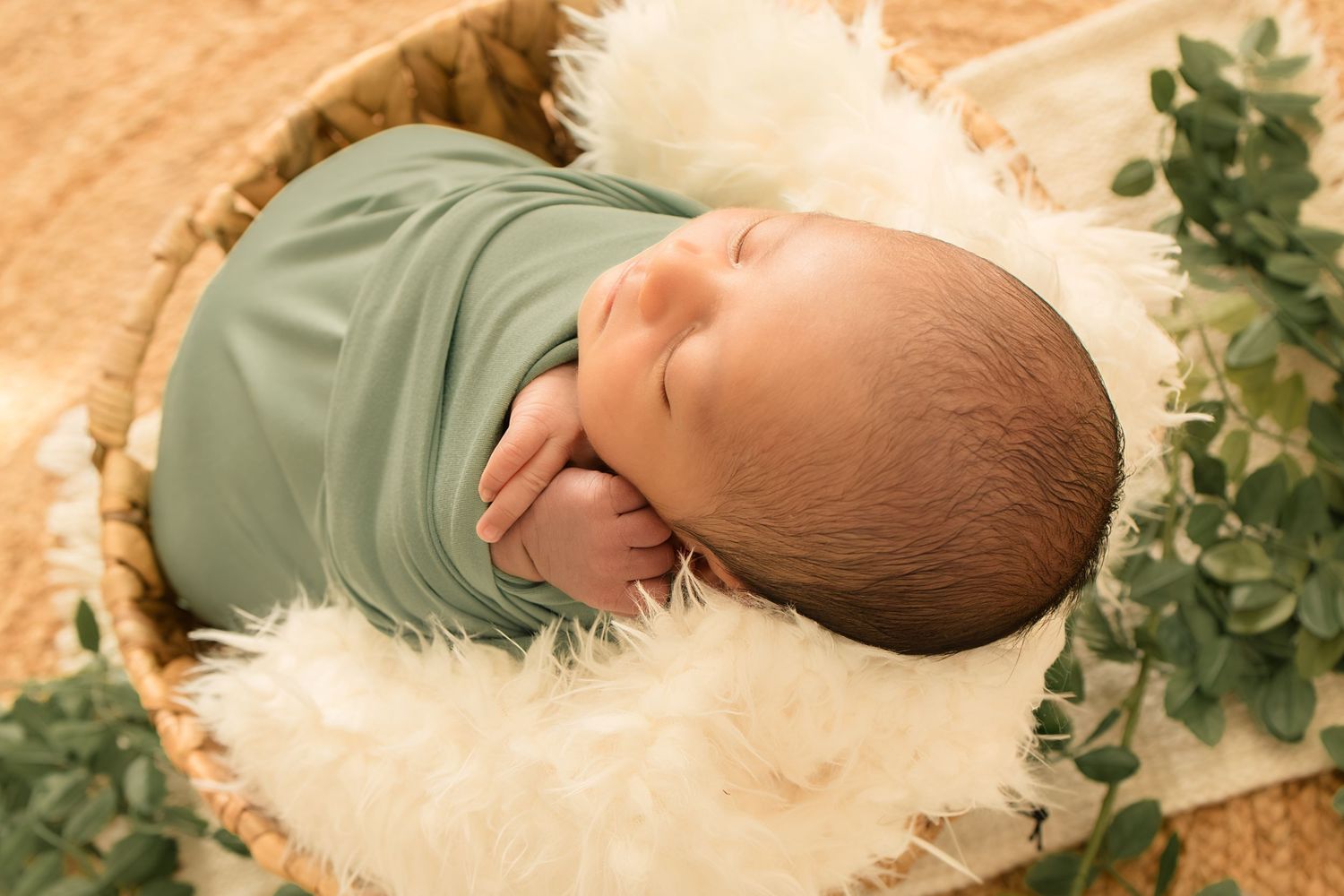 Newborn baby swaddled in green wrap sleeping peacefully in a wicker basket with white fur and eucalyptus.