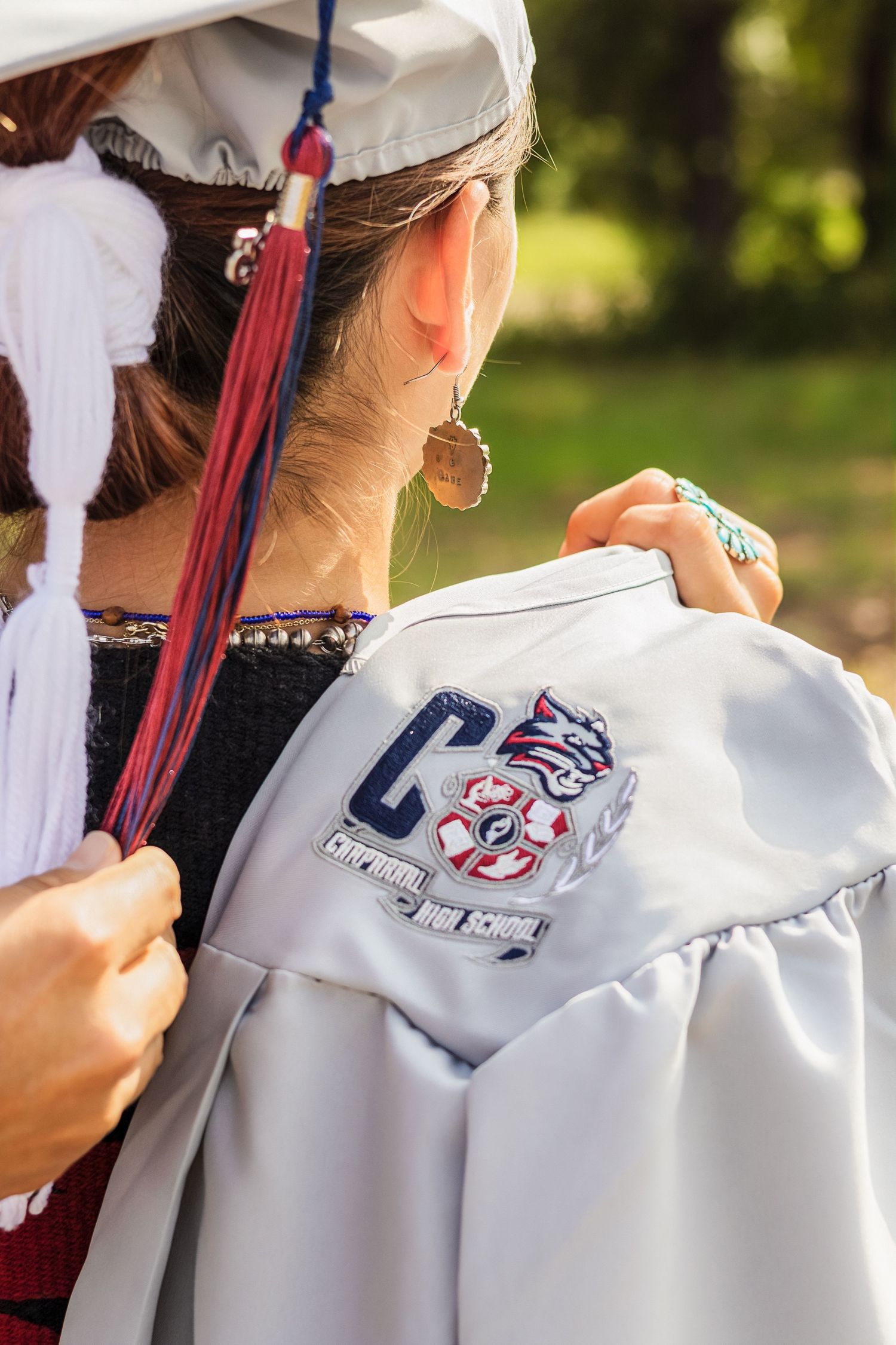 Close-up of a graduation gown with a high school emblem patch, worn by a student in a white cap with red tassel.