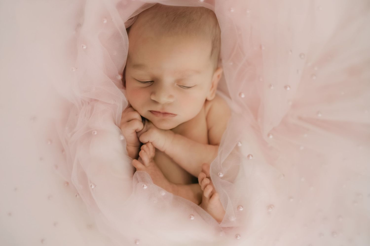 Sleeping newborn baby wrapped in soft pink tulle fabric with pearl details.