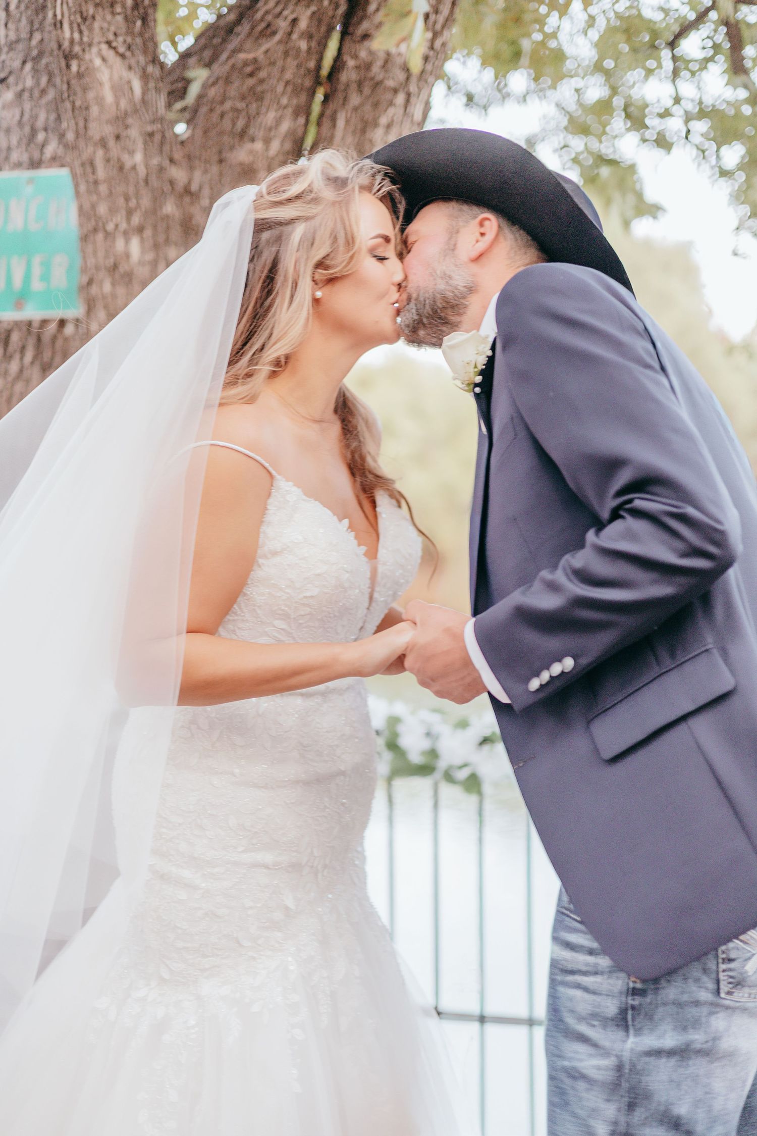Bride and groom share first kiss at outdoor wedding, groom wearing black cowboy hat and gray suit.