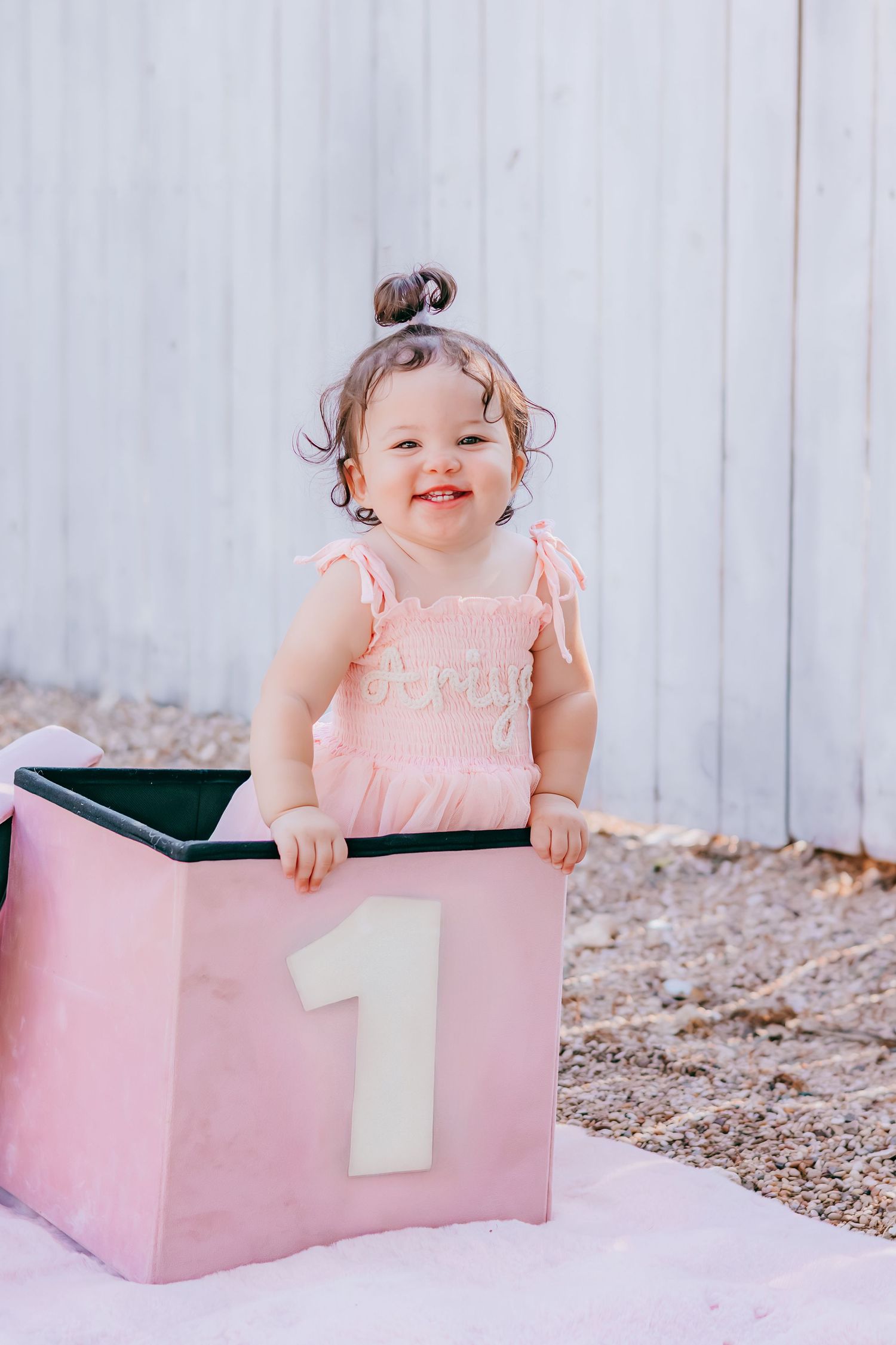 Smiling baby girl in pink romper standing in a pink box with number 1, celebrating her first birthday outdoors.