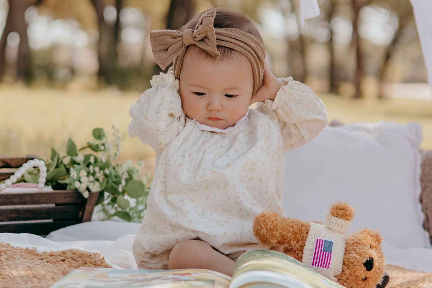 A baby girl in a white lace dress and tan bow headband sits on a blanket outdoors near a teddy bear and flowers.