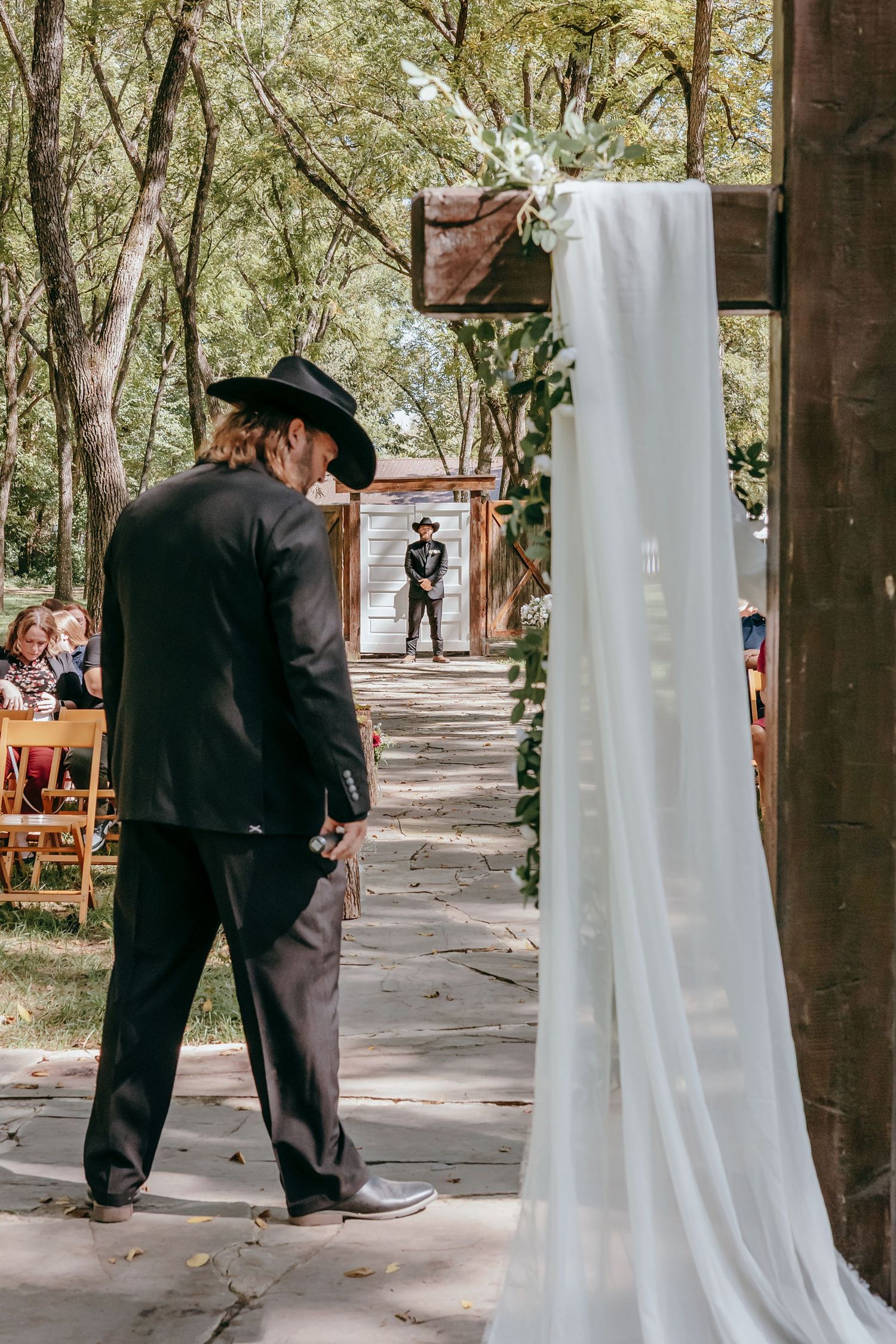 Groom in black cowboy hat waits at altar as young ring bearer walks down outdoor aisle.
