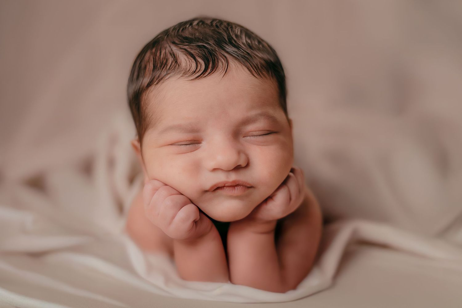 Peaceful newborn baby sleeping with hands on cheeks in classic froggy pose on beige backdrop.