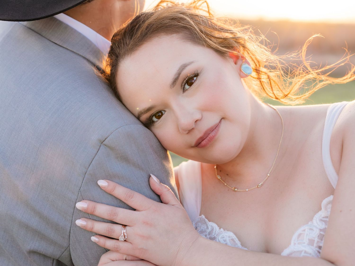 Bride rests her head on groom's shoulder, showcasing her wedding ring in a golden sunset portrait.