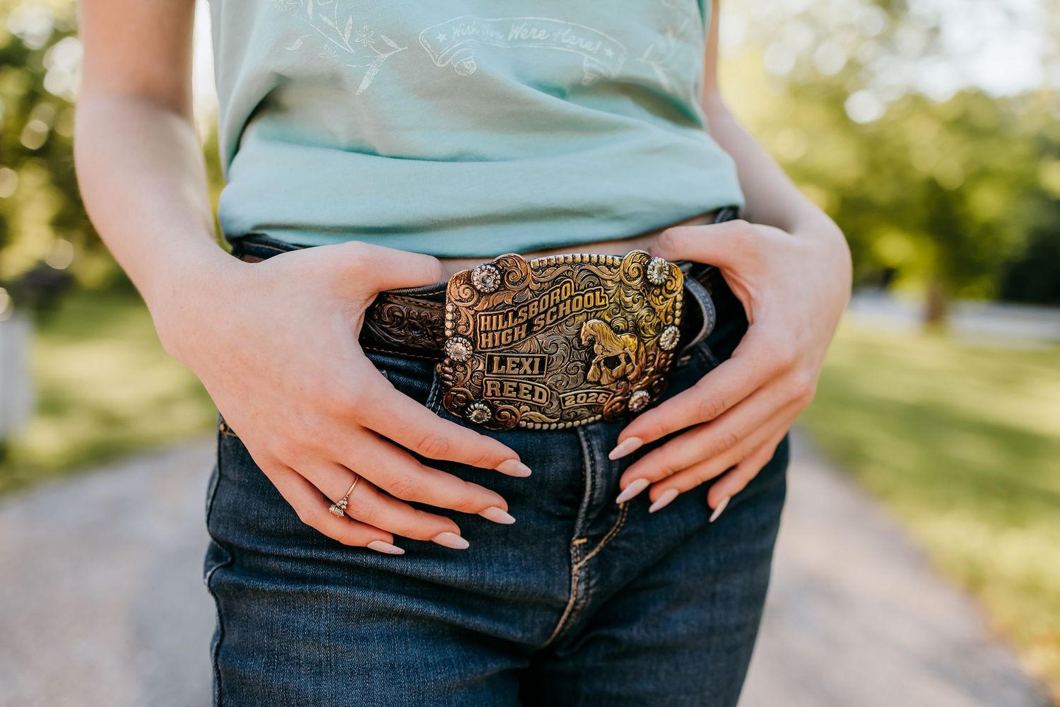Close-up of a woman wearing jeans with a decorative Western gold belt buckle, showing manicured dark nails.