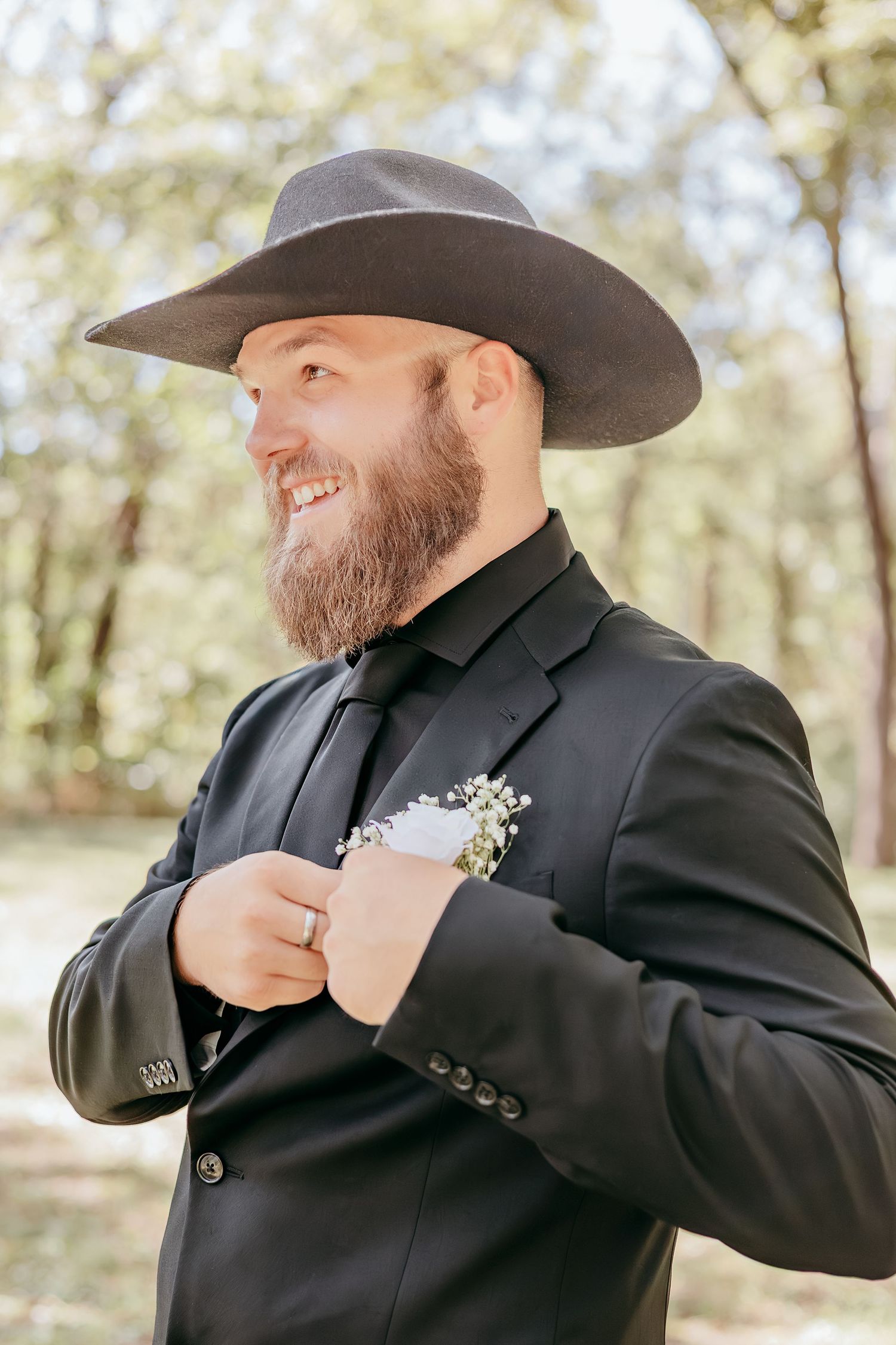 Bearded groom in black cowboy hat and suit with boutonniere smiling outdoors in wooded setting.