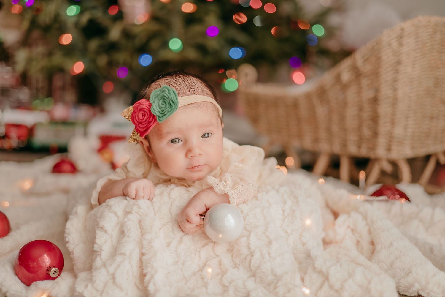 Awake newborn baby wrapped in cream blanket with Christmas ornaments and festive tree in background.