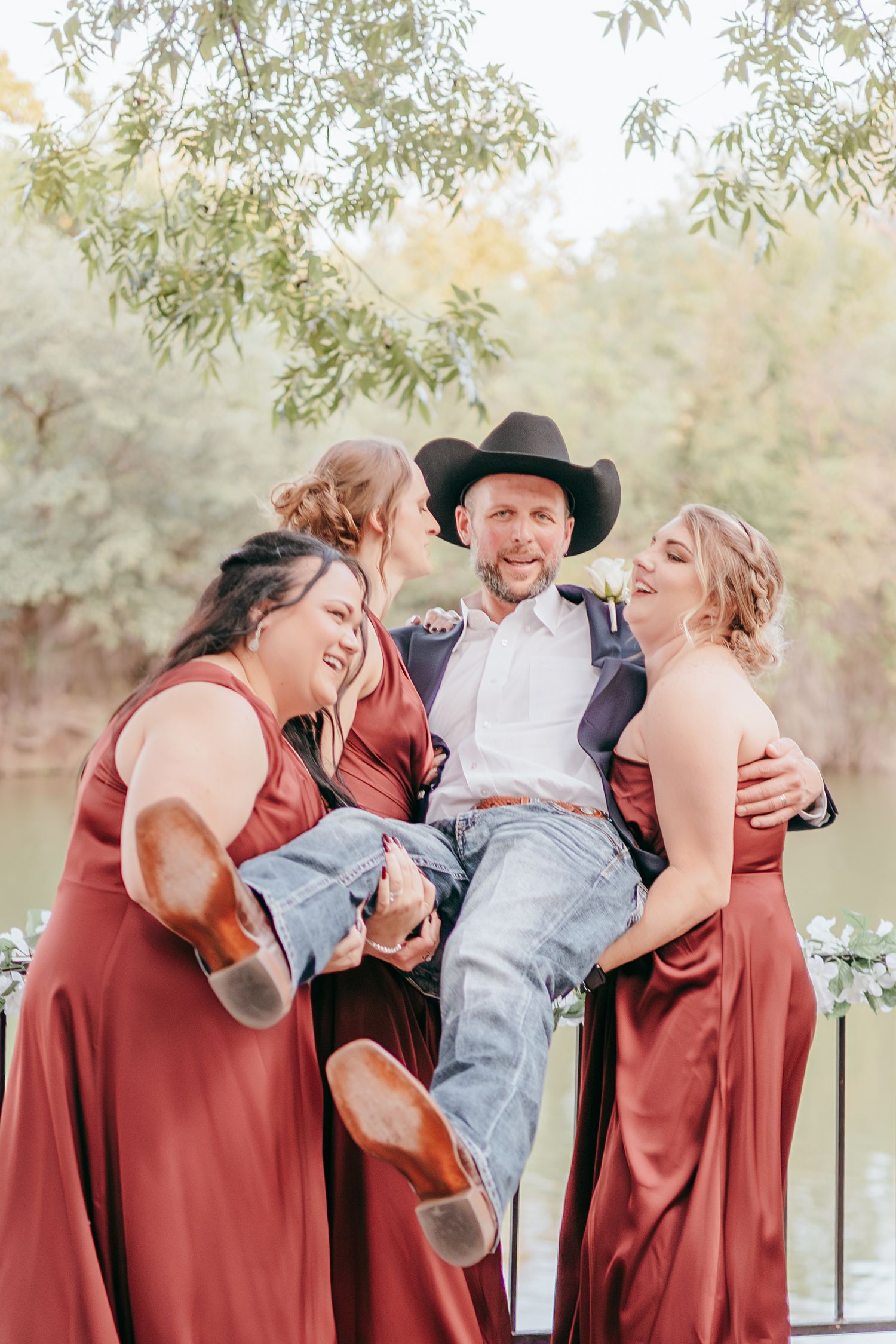 Three bridesmaids in rust satin dresses playfully lift groom in cowboy hat and jeans at outdoor wedding venue.
