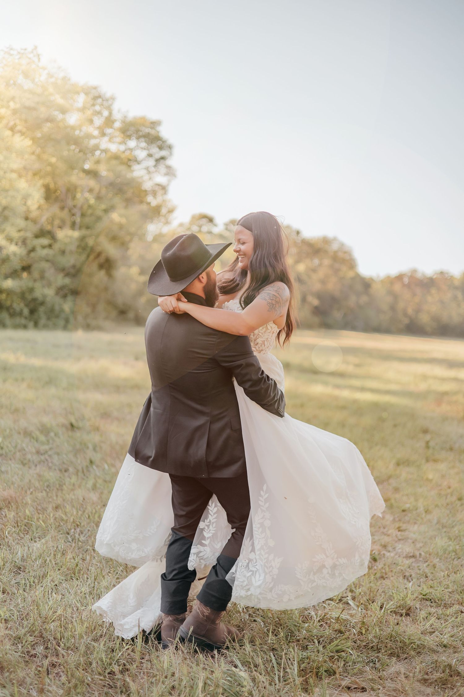Bride and groom dancing joyfully in open field at sunset, groom in cowboy hat and black suit.