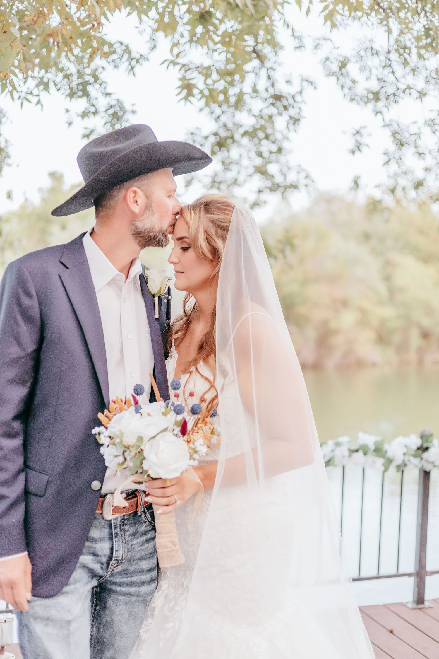 Groom in cowboy hat and bride with veil holding bouquet near waterfront trees.
