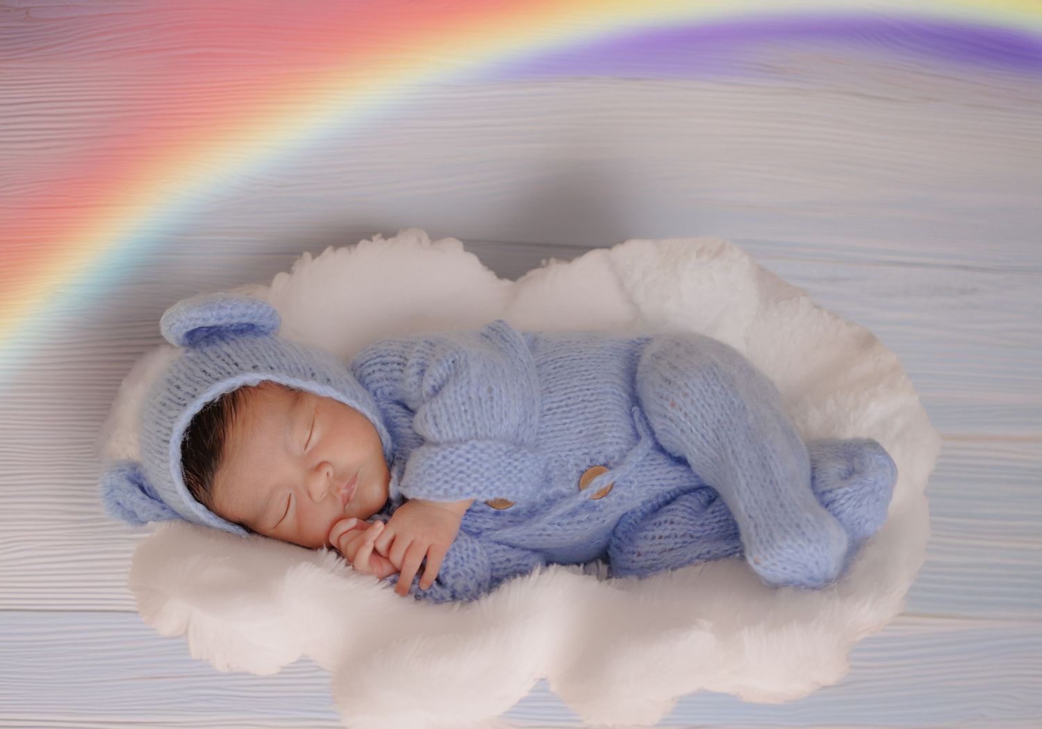 Sleeping newborn baby in blue knit outfit on cloud prop with rainbow background.