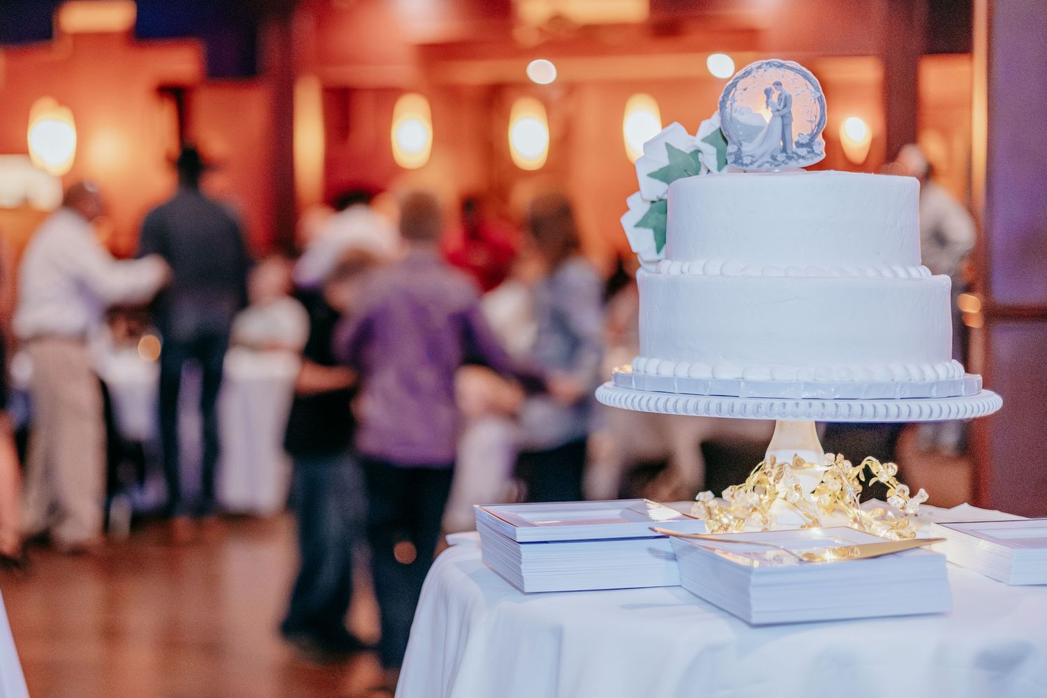 Elegant two-tier white wedding cake with floral topper displayed on table, guests dancing in blurred reception hall background.