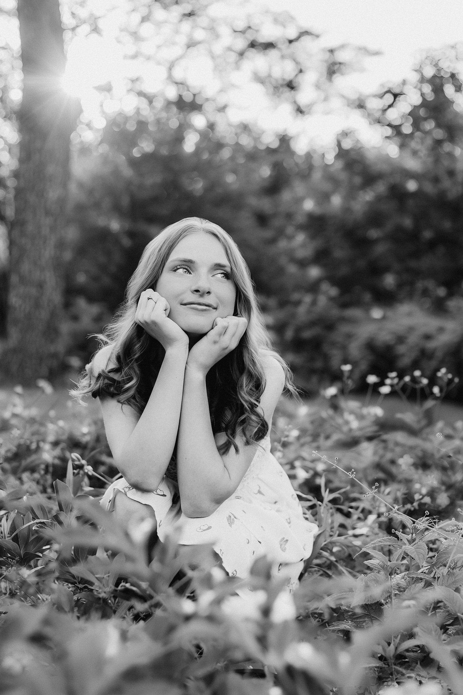 Black and white portrait of a young woman sitting in a garden, resting her face in her hands, gazing upward dreamily.