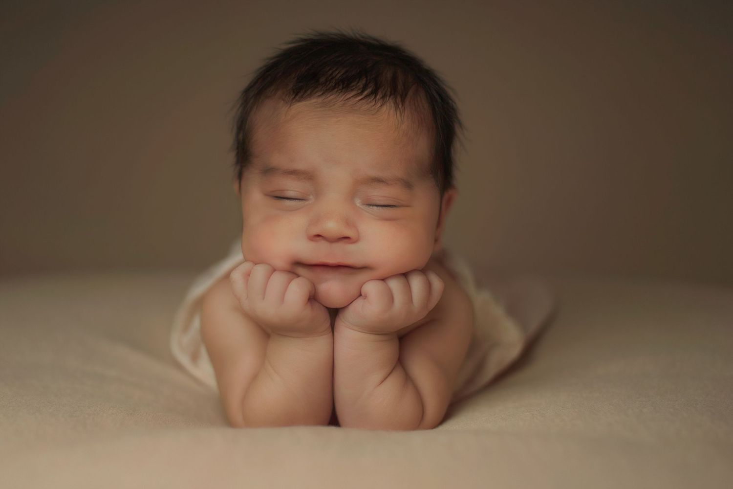 Peaceful newborn baby sleeping with chin resting on tiny fists, wrapped in white fabric on a soft cream blanket.