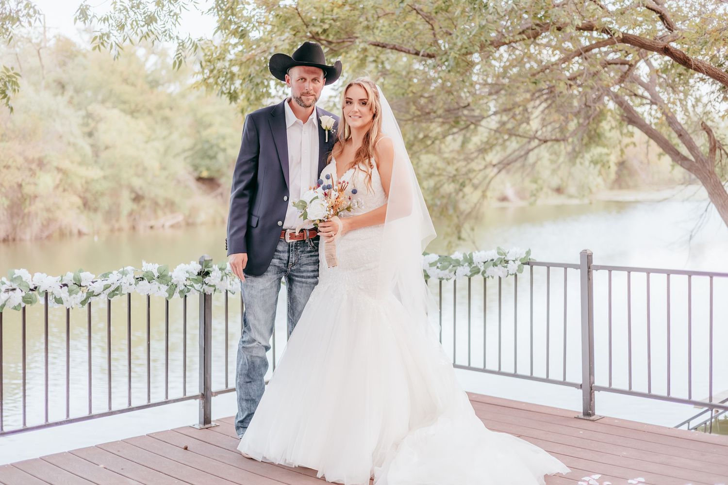 Bride in white gown and groom in black blazer with cowboy hat pose on a wooden deck by a river.