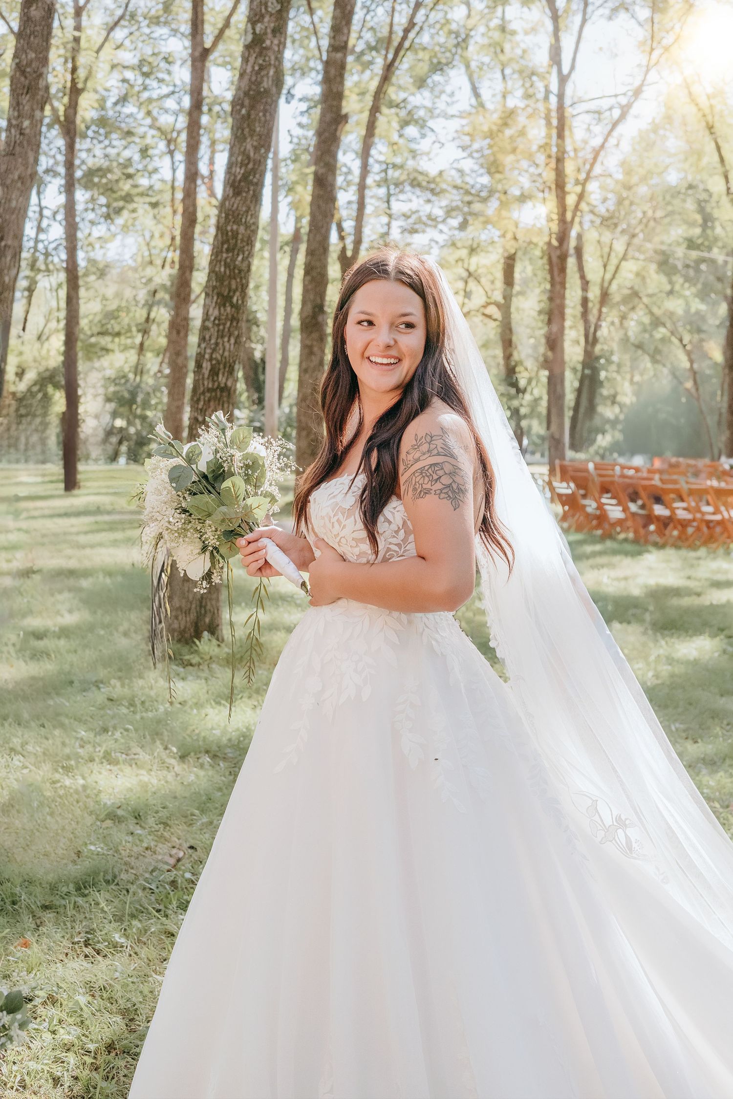 Bride in lace ballgown with veil and bouquet smiling in sunlit forest wedding venue.