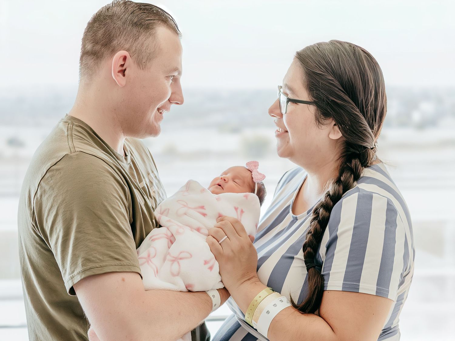 Happy new parents smiling and holding their swaddled newborn baby together in a hospital room.