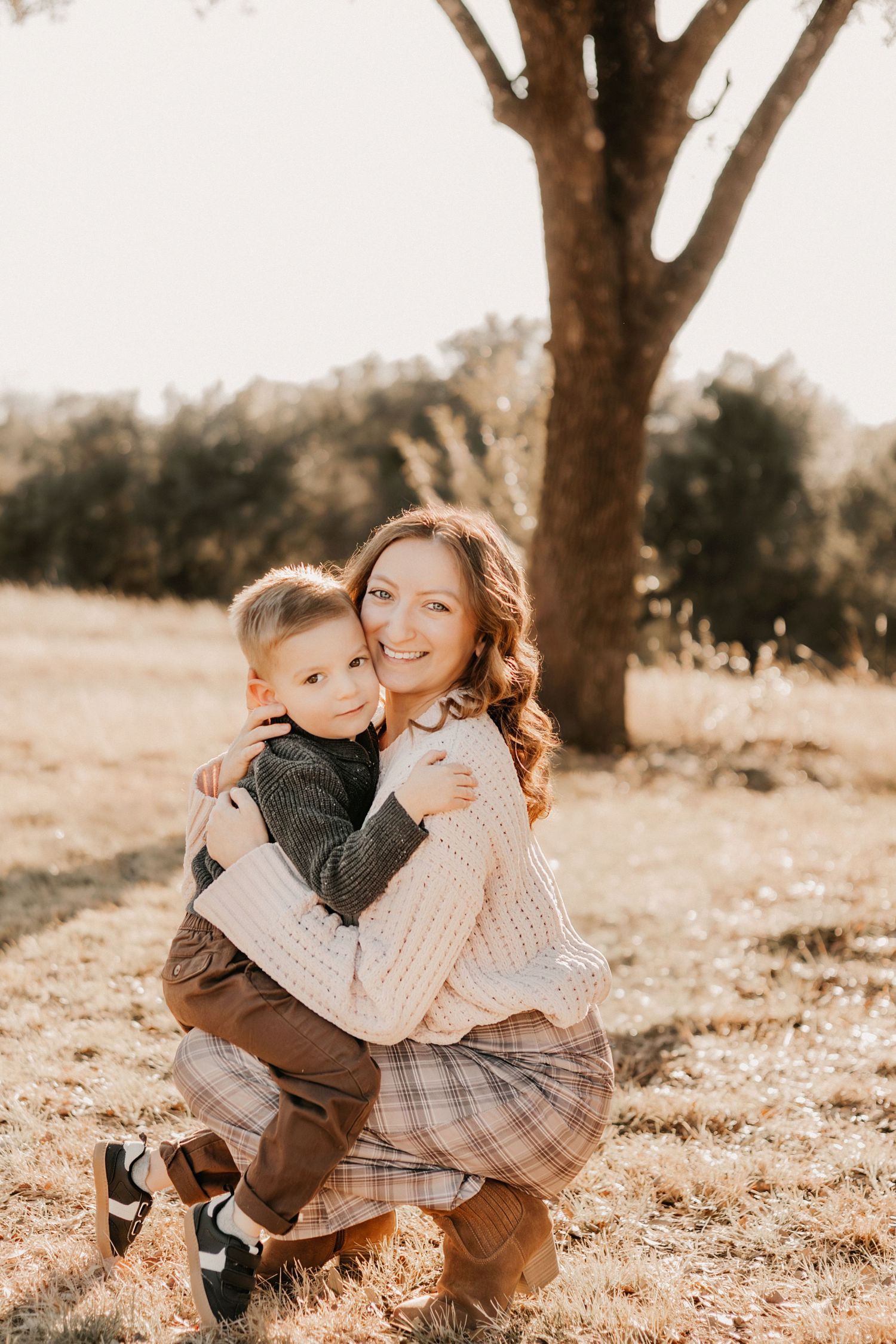 A happy mother in a plaid skirt hugging her smiling toddler son outdoors near a tree.