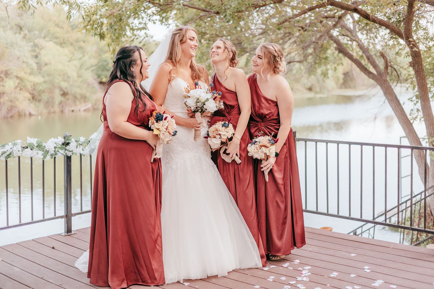 Bride in white gown with three bridesmaids in rust-red dresses holding bouquets on a wooden deck by a river.