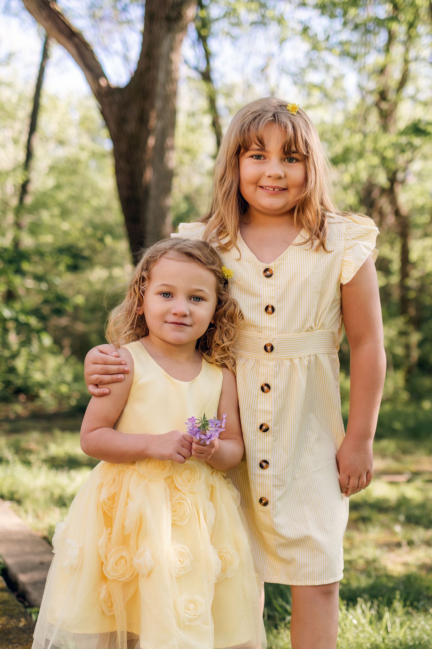 Two young sisters in matching yellow dresses pose together outdoors in a sunny green park, with the older girl hugging the younger one.