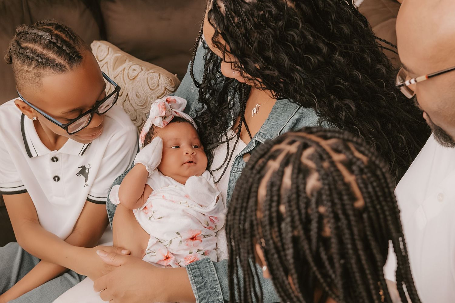 Family gathered indoors admiring newborn baby in floral swaddle with bow headband on couch.