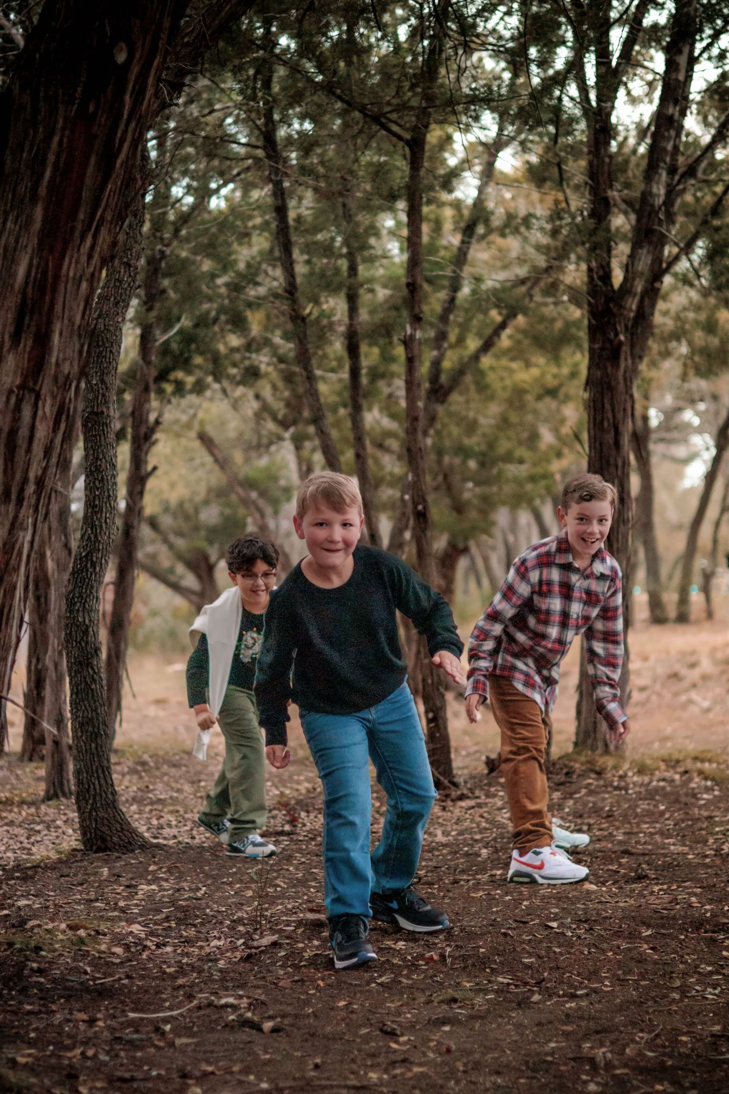 Three young boys run and play together along a wooded forest path in casual outdoor clothing.
