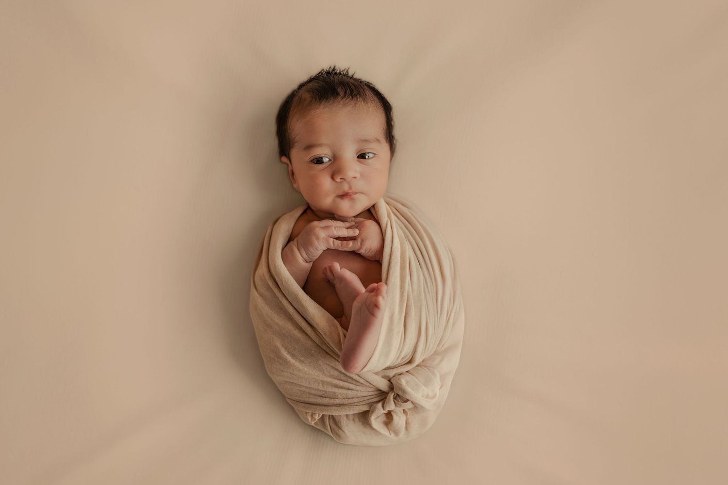 Newborn baby wrapped snugly in a cream swaddle with hands near face on a beige background.