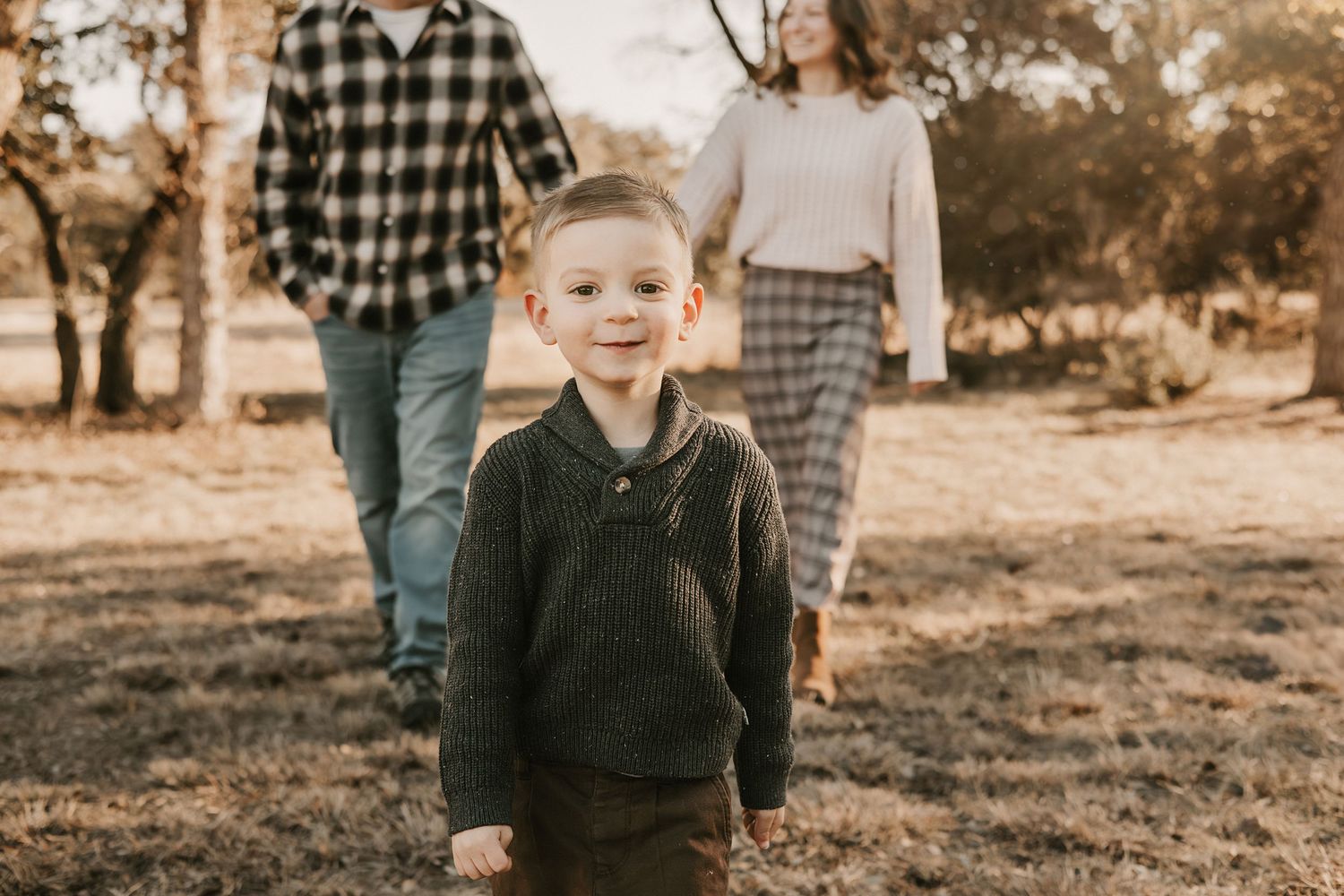 A smiling toddler boy walks ahead of his parents through a golden tree-lined field.