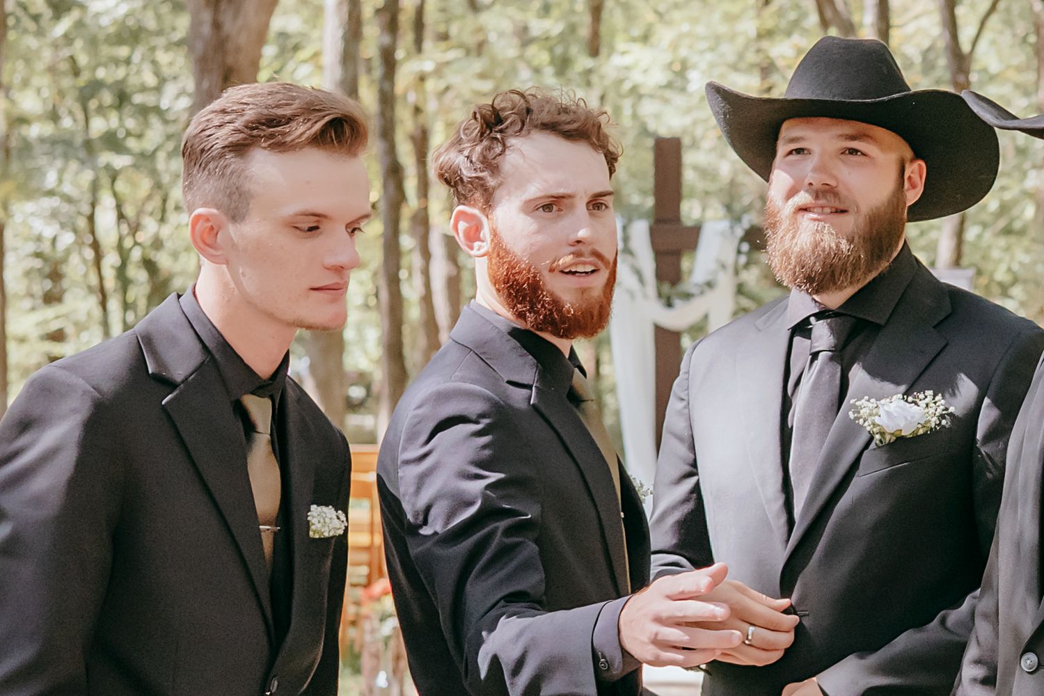 Three groomsmen in dark suits with boutonnieres conversing outdoors in wooded wedding setting on sunny day.