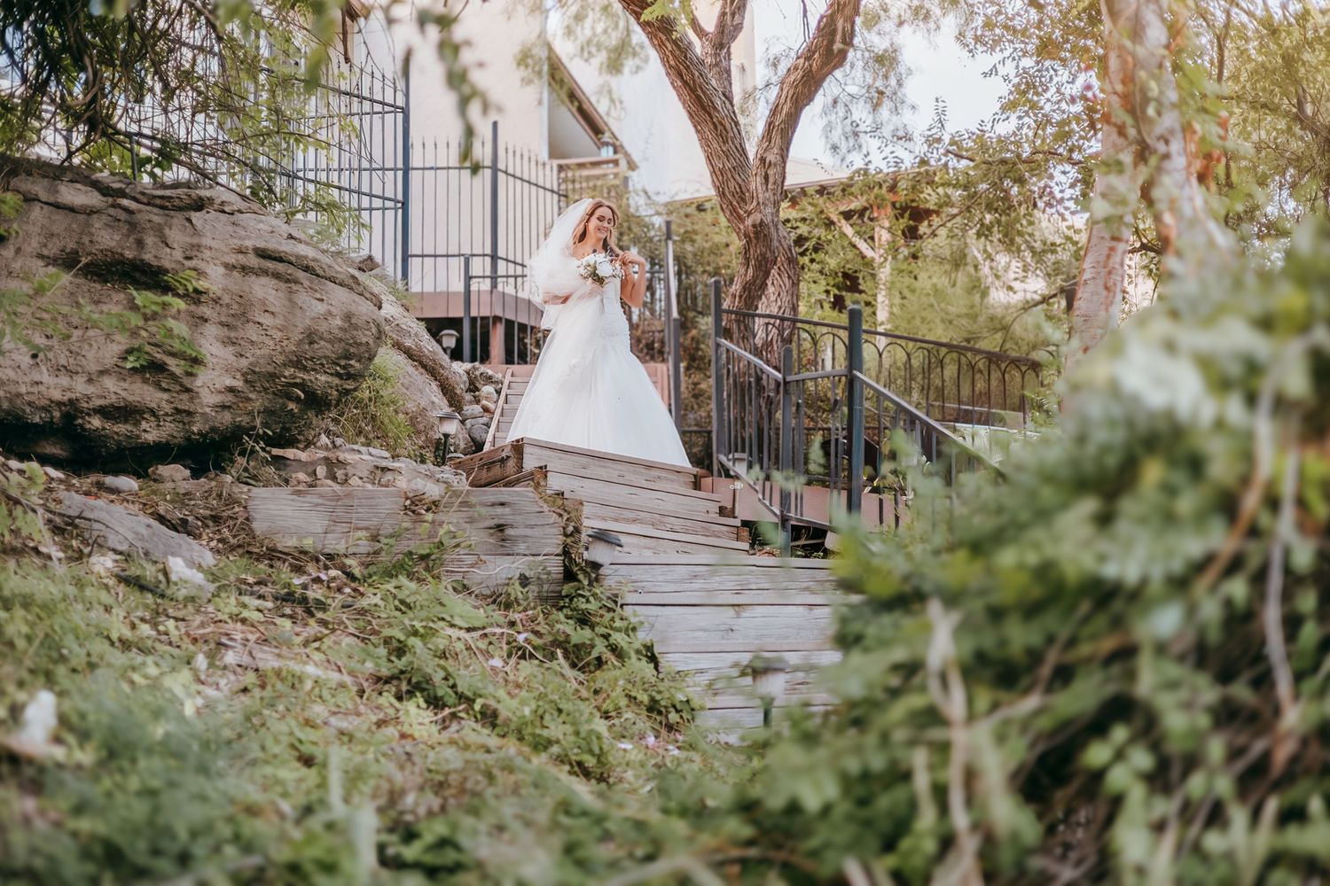 Bride in white gown descending outdoor stone steps surrounded by lush greenery.
