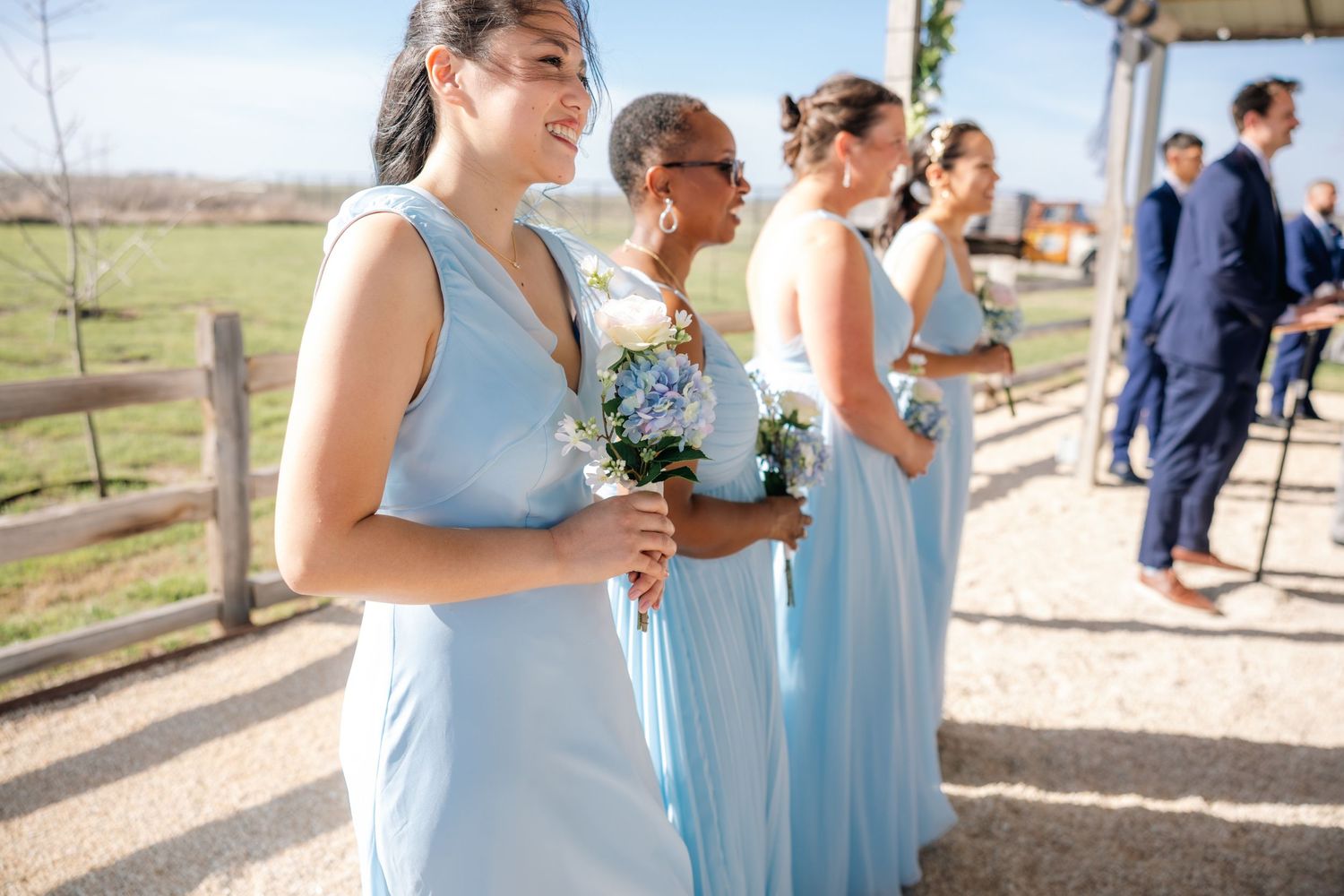 Bridesmaids in light blue gowns holding floral bouquets smile during an outdoor rustic wedding ceremony.