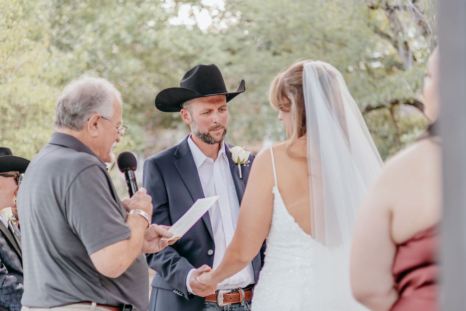 Outdoor wedding ceremony with groom in cowboy hat and blue suit exchanging vows with bride in white dress and veil.