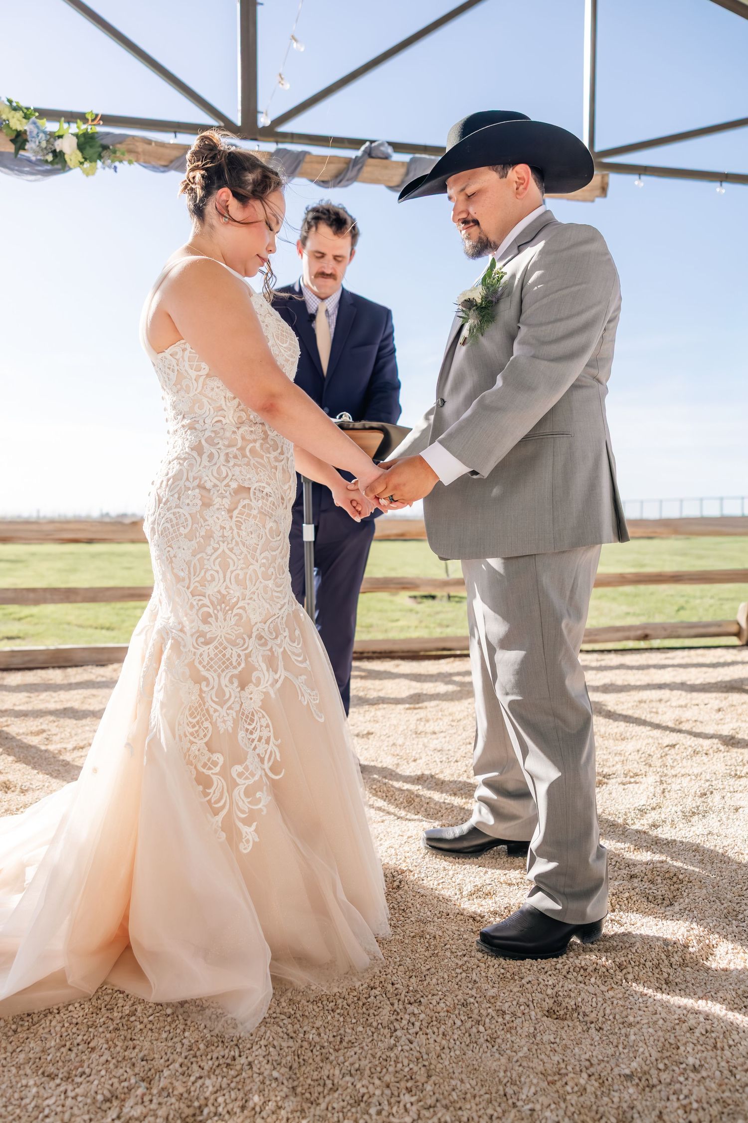Bride and groom hold hands during outdoor wedding ceremony while officiant stands behind them under a canopy.