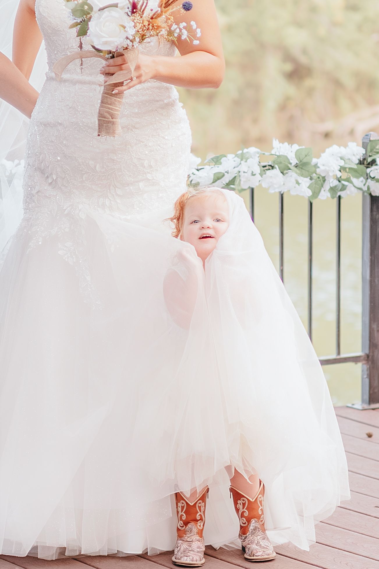 A toddler flower girl in a white tulle dress and cowboy boots peeks out from behind a bride's wedding gown.