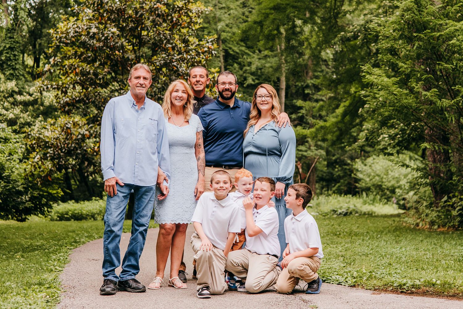 Family of eight poses on a garden path surrounded by lush green trees for an outdoor portrait.