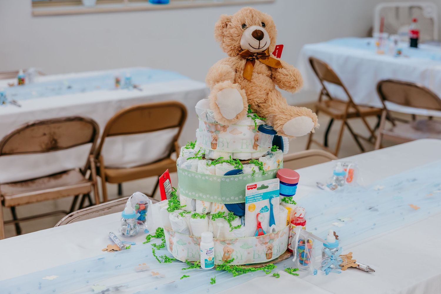 A tiered diaper cake topped with a teddy bear displayed on a table at a baby shower party.