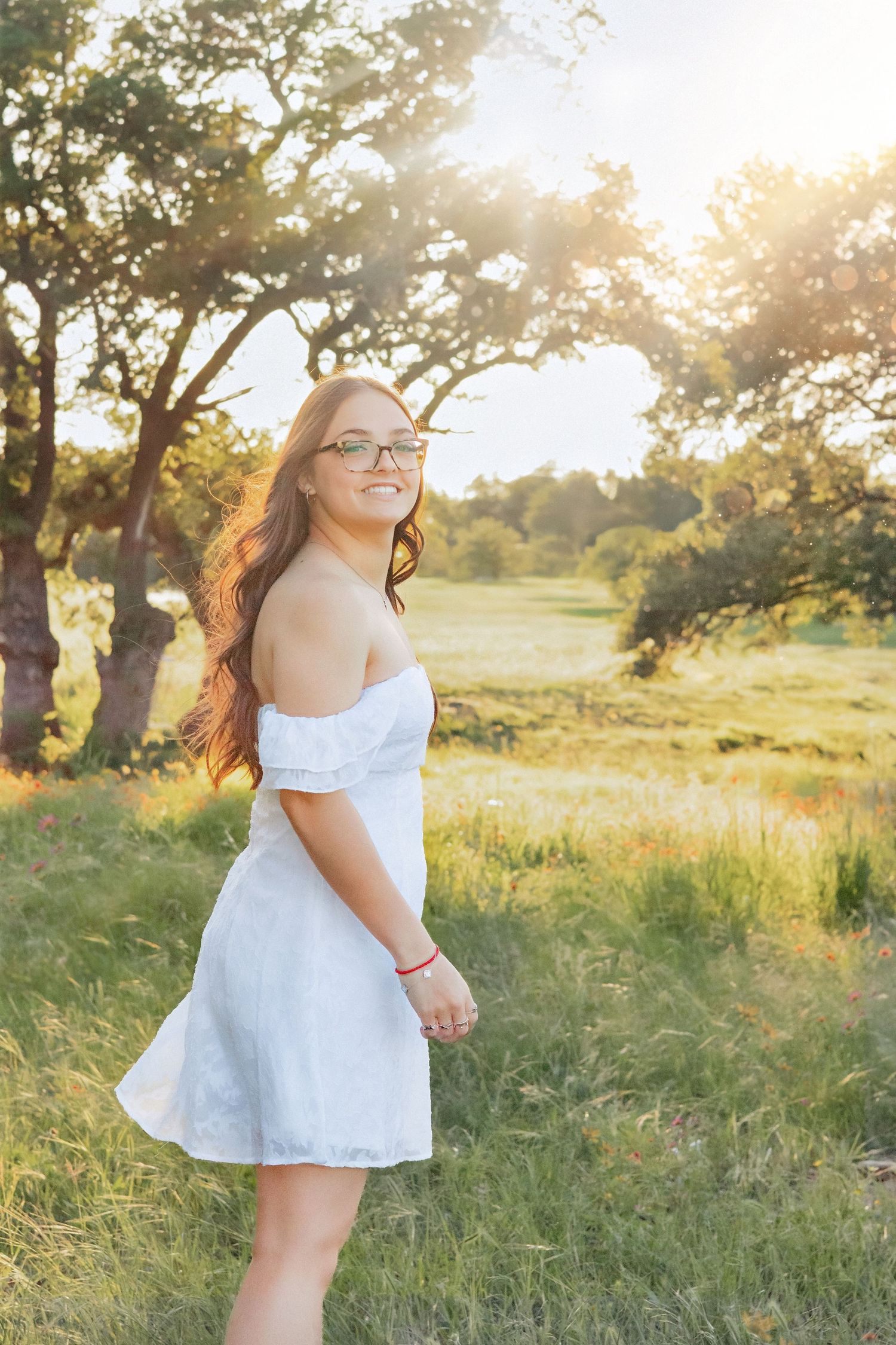Young woman in white off-shoulder dress smiling in sunlit meadow with trees during golden hour.