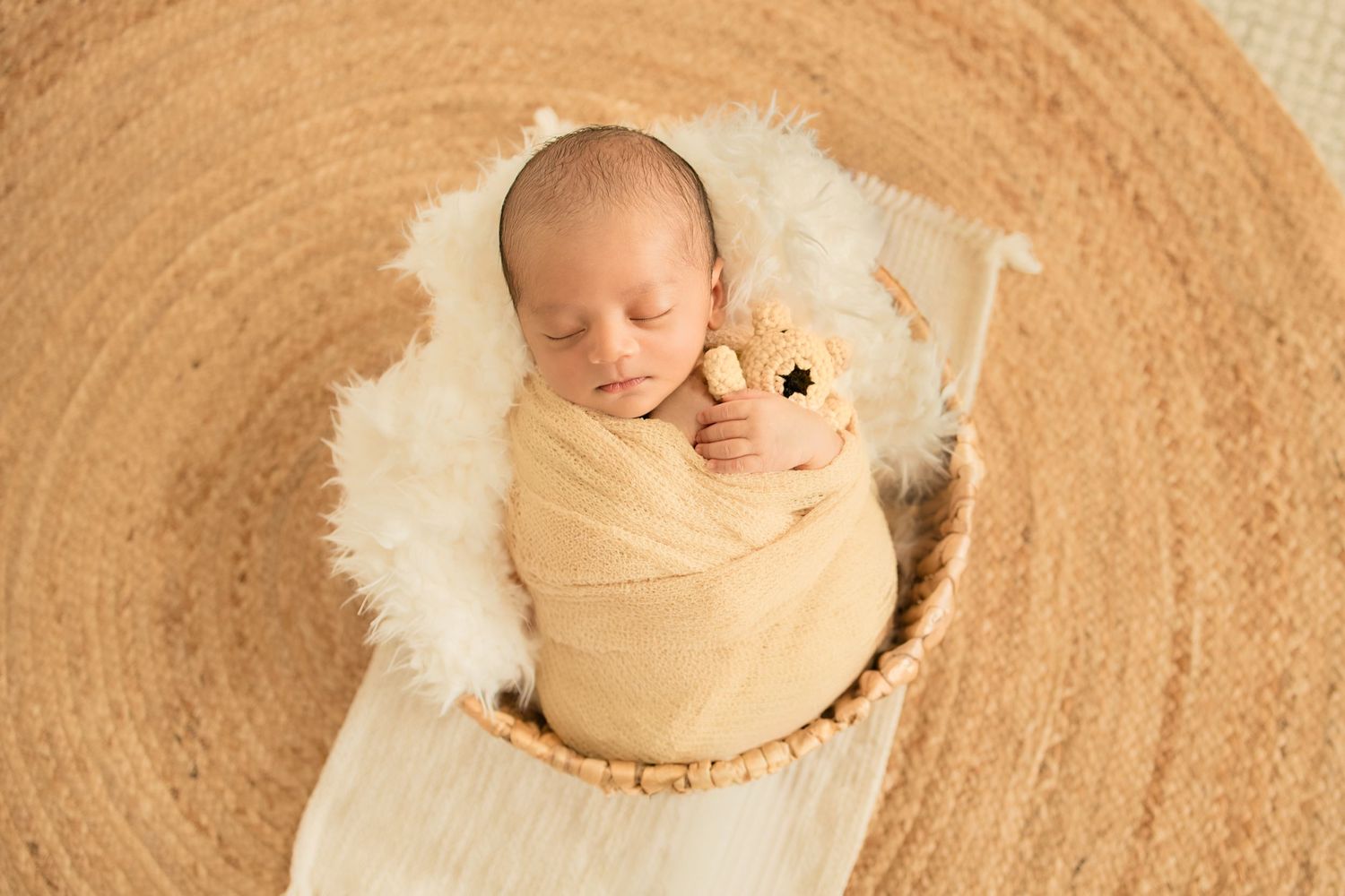 Newborn baby swaddled in yellow wrap holding a small stuffed animal, sleeping peacefully in a wicker basket.