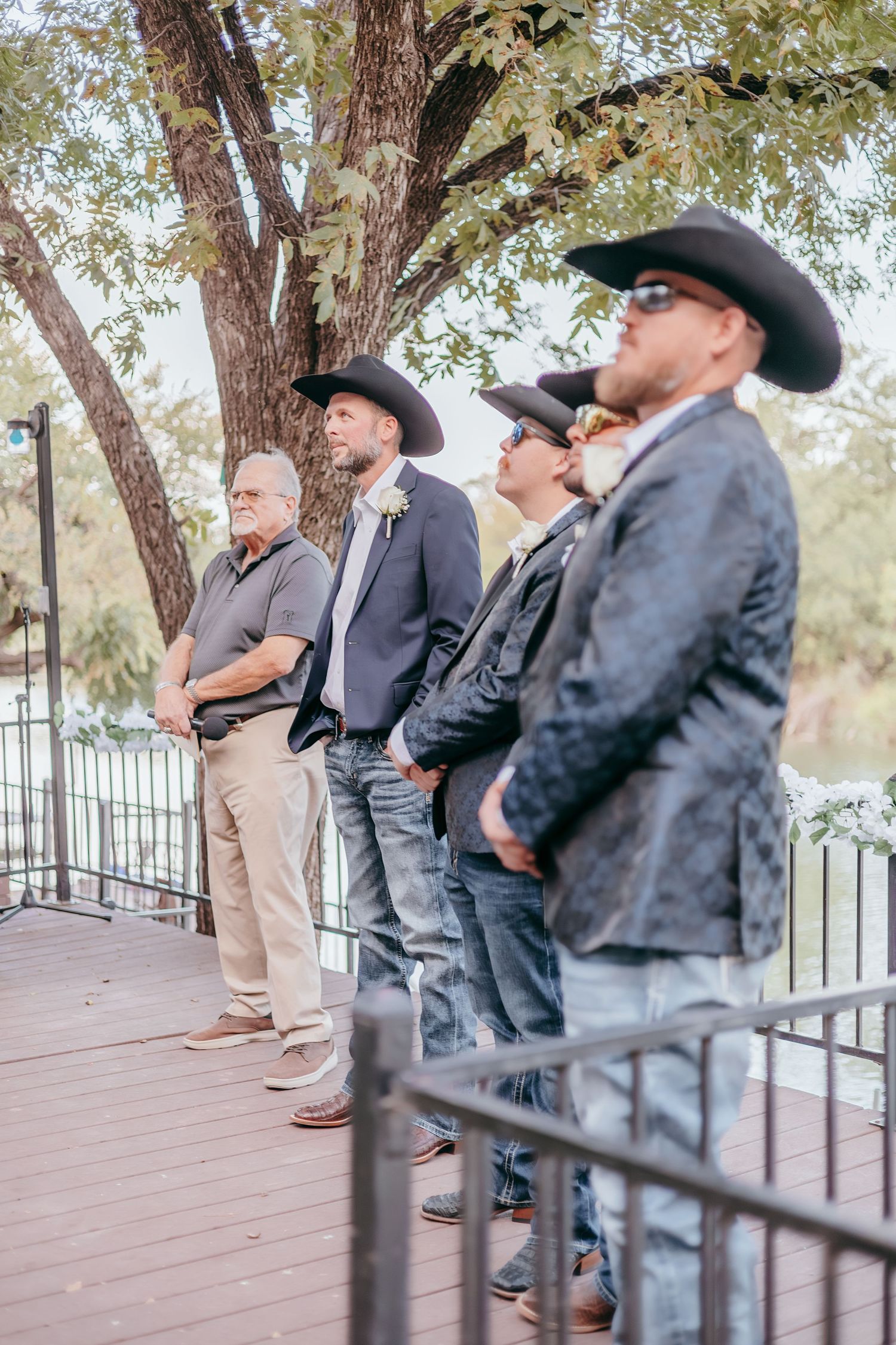Groomsmen in black cowboy hats and blazers standing on wooden deck during outdoor wedding ceremony.