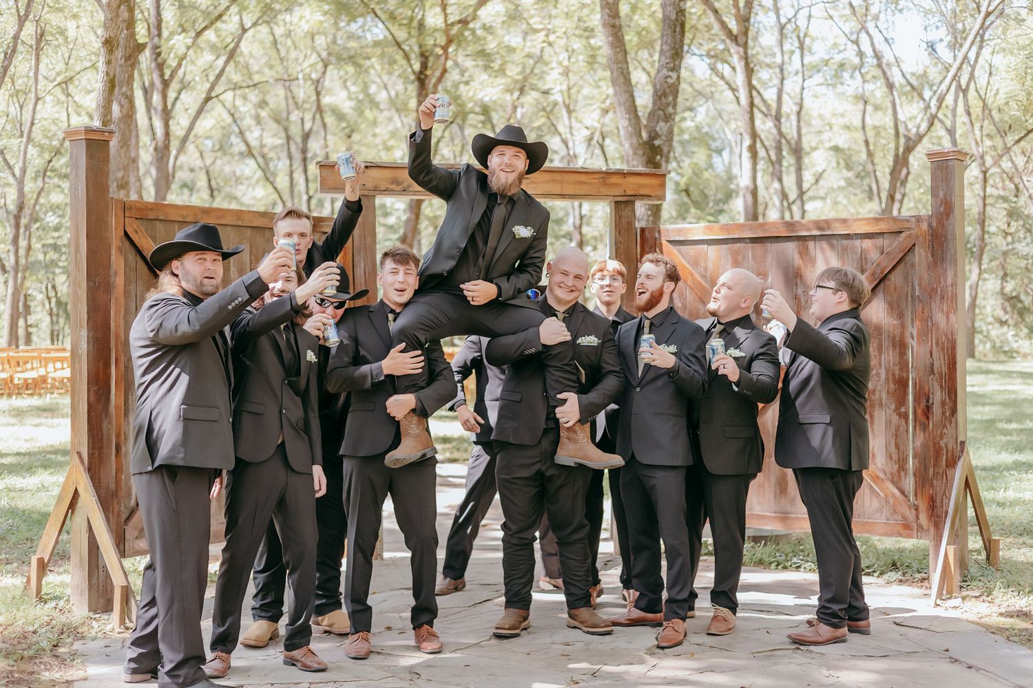 Groomsmen in black suits lifting groom in cowboy hat outdoors at rustic wedding venue surrounded by trees.