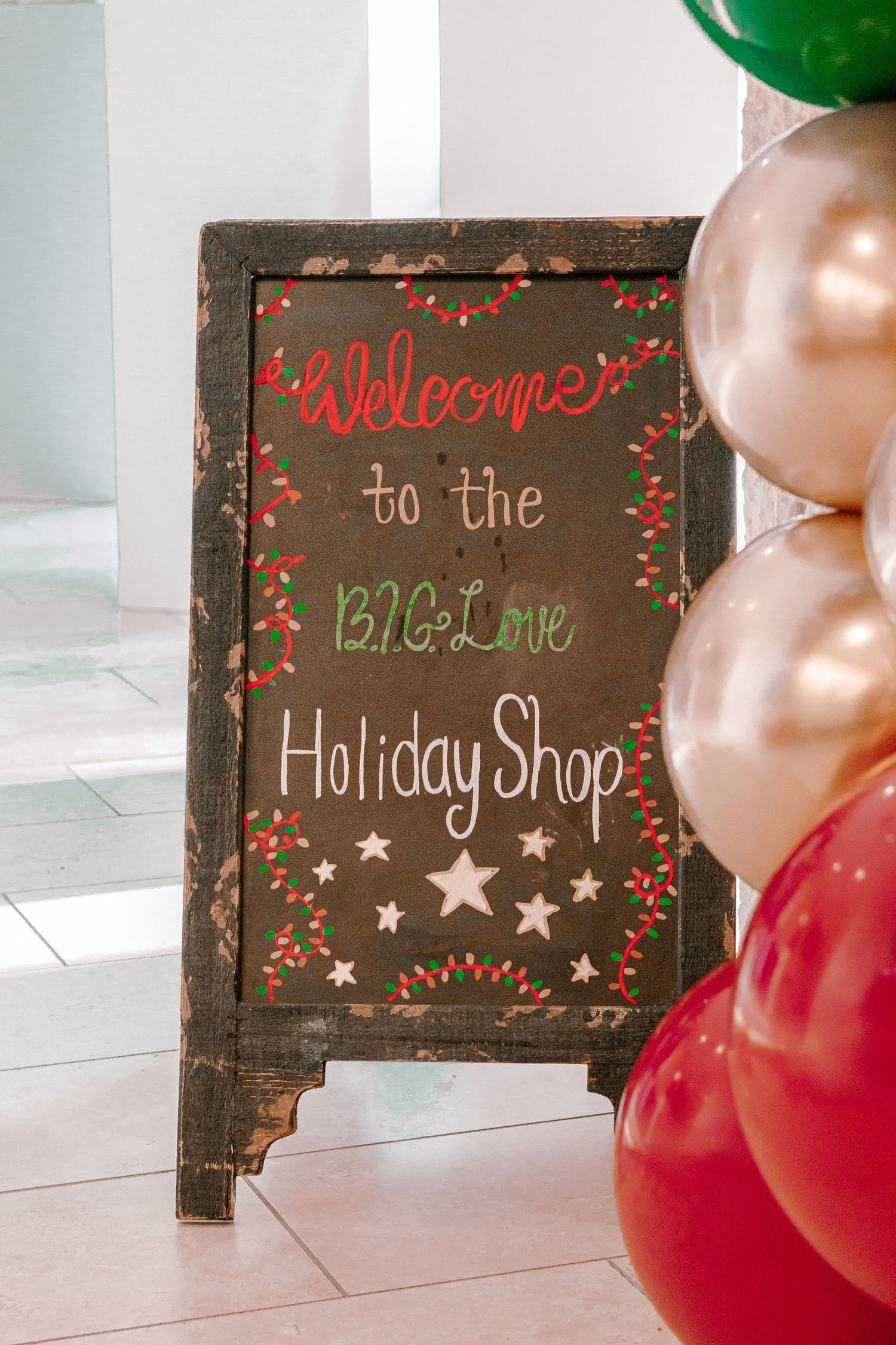 Chalkboard welcome sign for the BIG Love Holiday Shop decorated with stars and holly, surrounded by festive red, green, and gold balloons.