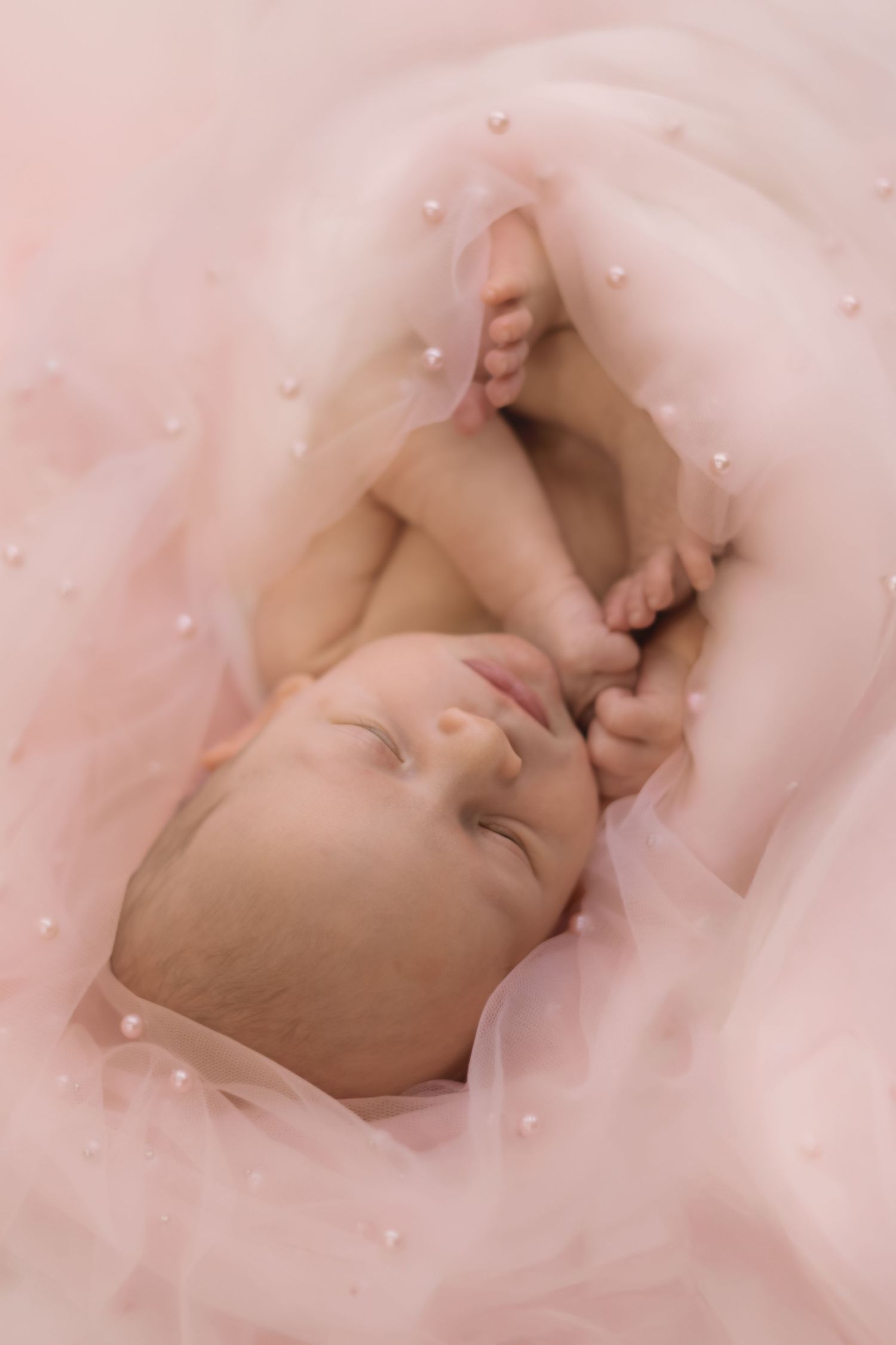 Newborn baby lying on soft pink tulle fabric with pearl embellishments all around.