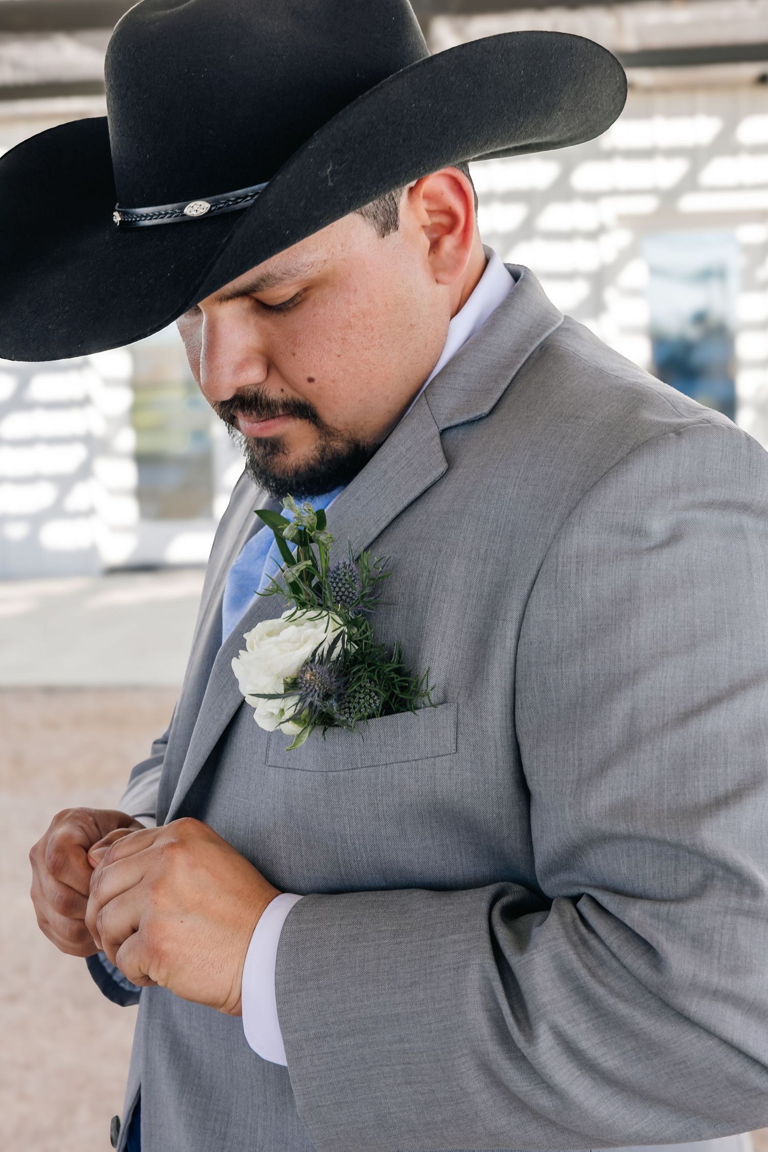 Groom in black cowboy hat and gray suit adjusts his white rose and thistle boutonniere before the ceremony.