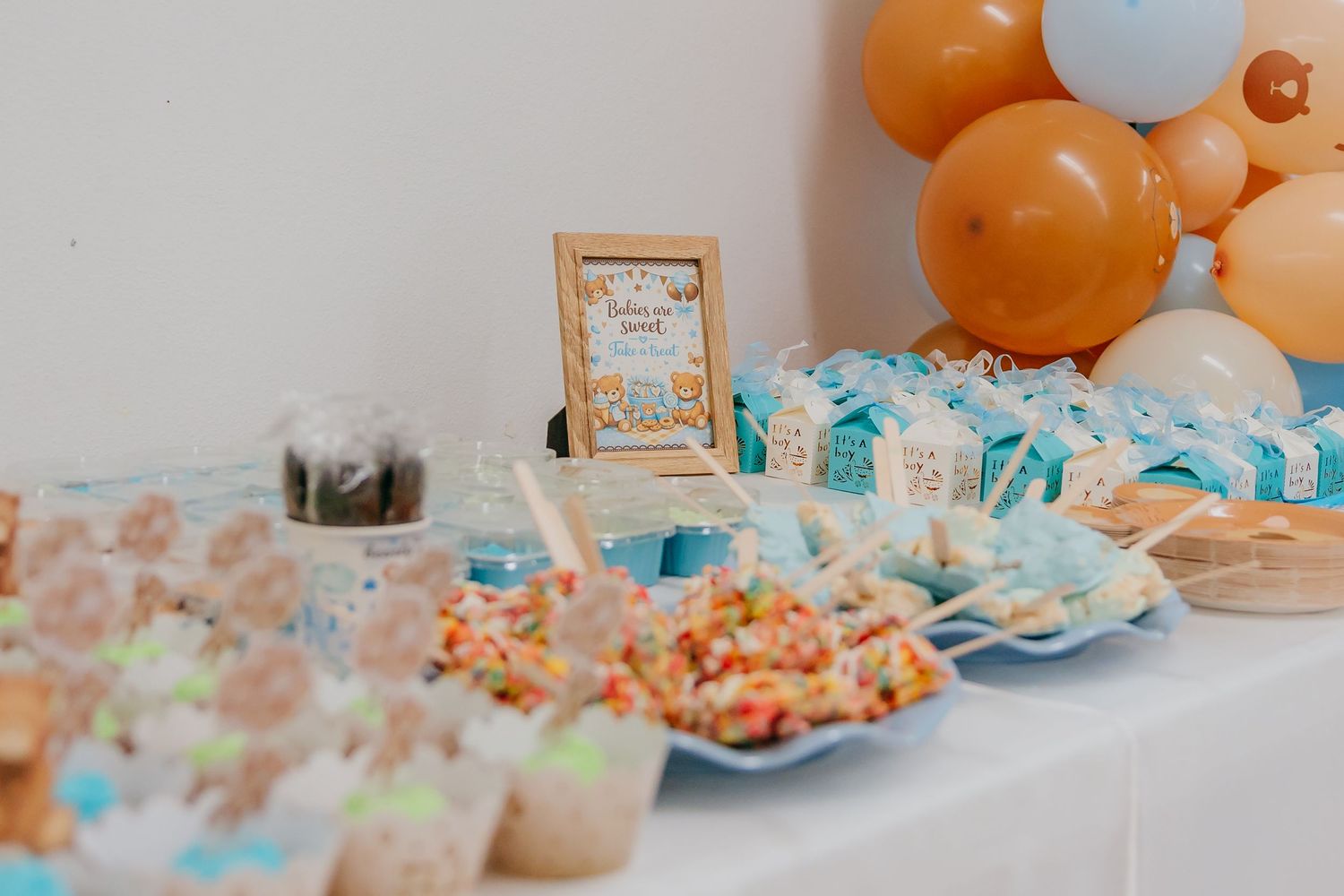 Colorful dessert table with treats, blue gift boxes, a framed sign, and orange balloon garland for a celebration.