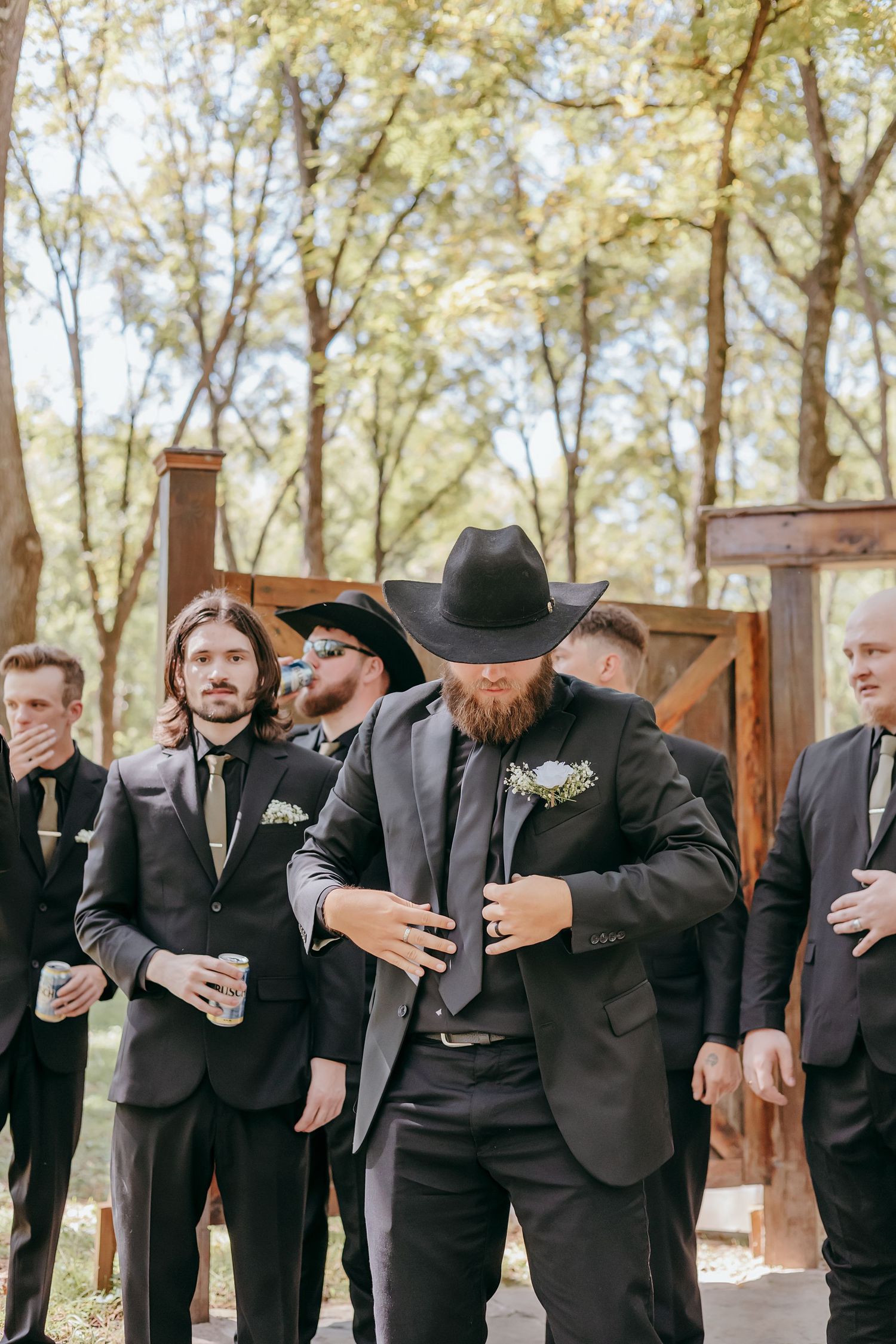 Groom in black suit and cowboy hat adjusts jacket surrounded by groomsmen in matching black suits outdoors.