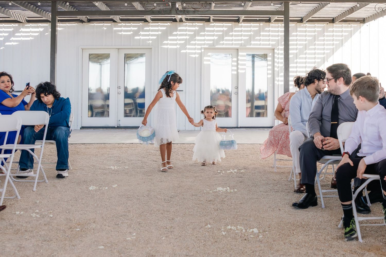 Two flower girls in white dresses with blue sashes walk down the wedding ceremony aisle.