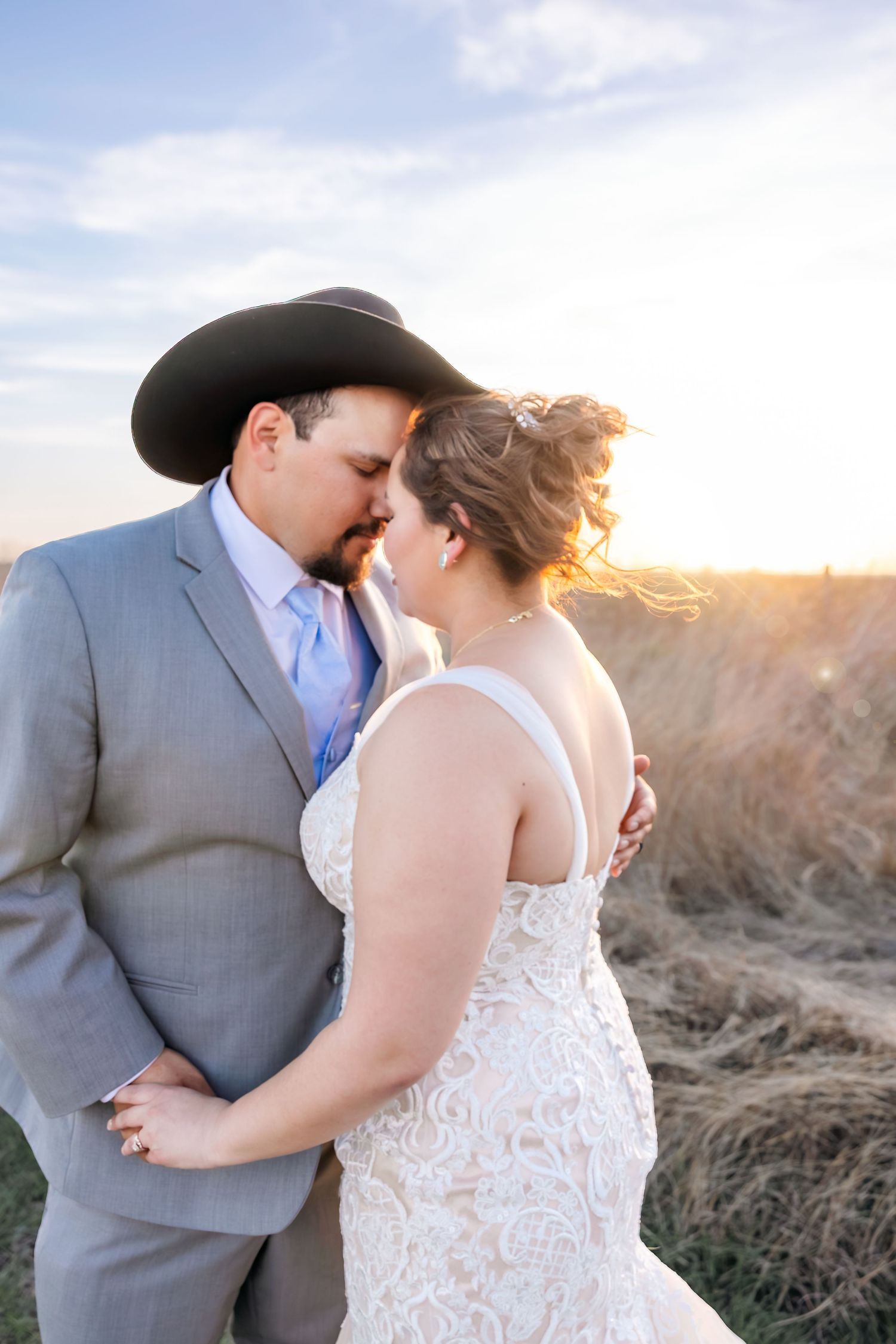 Bride and groom share a romantic embrace in a golden sunset field, groom wearing a black cowboy hat.