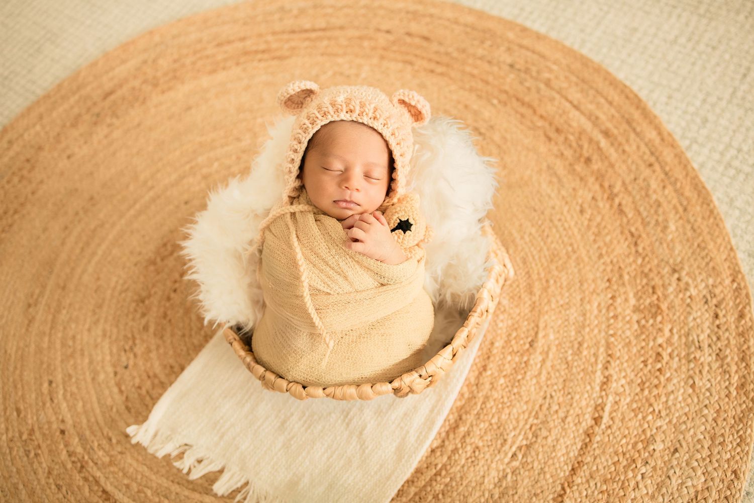 Sleeping newborn baby wrapped in cream swaddle wearing bear bonnet in wooden bowl on round jute rug.