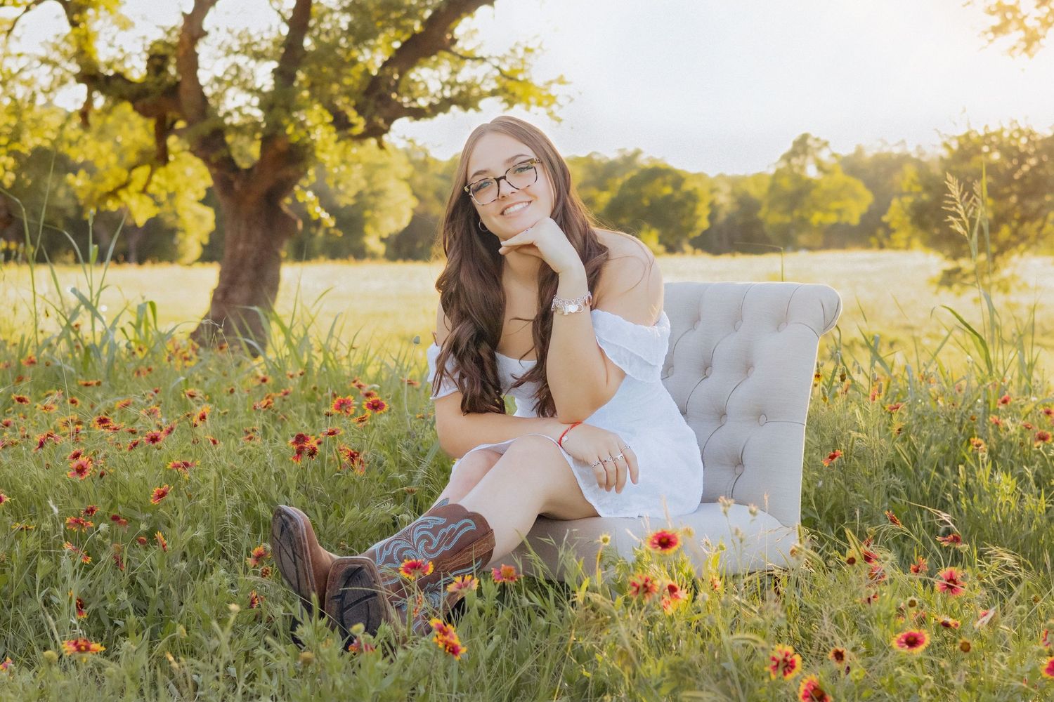 Young woman in white dress and cowboy boots sits in a tufted chair among wildflowers in a sunny field.